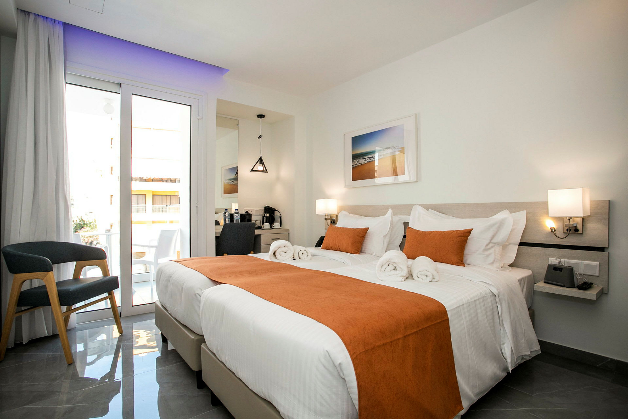 Ett ljust hotellrum med två enkelsängar som är bäddade med vita lakan och orangea dekorationer. Det finns en balkong med glasdörrar, en stol och ett skrivbord med spegel. (AI-genererad text)