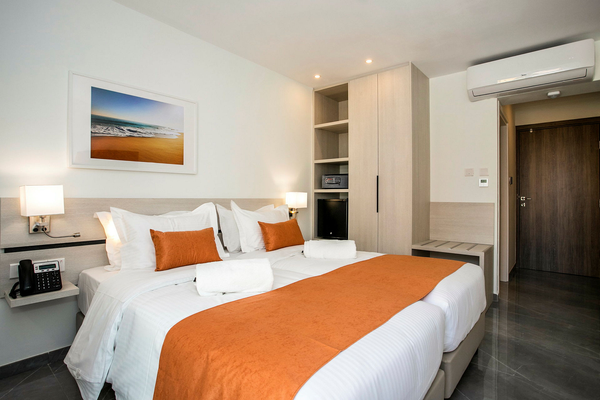 Ett hotellrum med en dubbelsäng täckt av vit och orange sängklädsel. Rummet har moderna möbler, luftkonditionering och väggkonst ovanför sängen. (AI-genererad text)