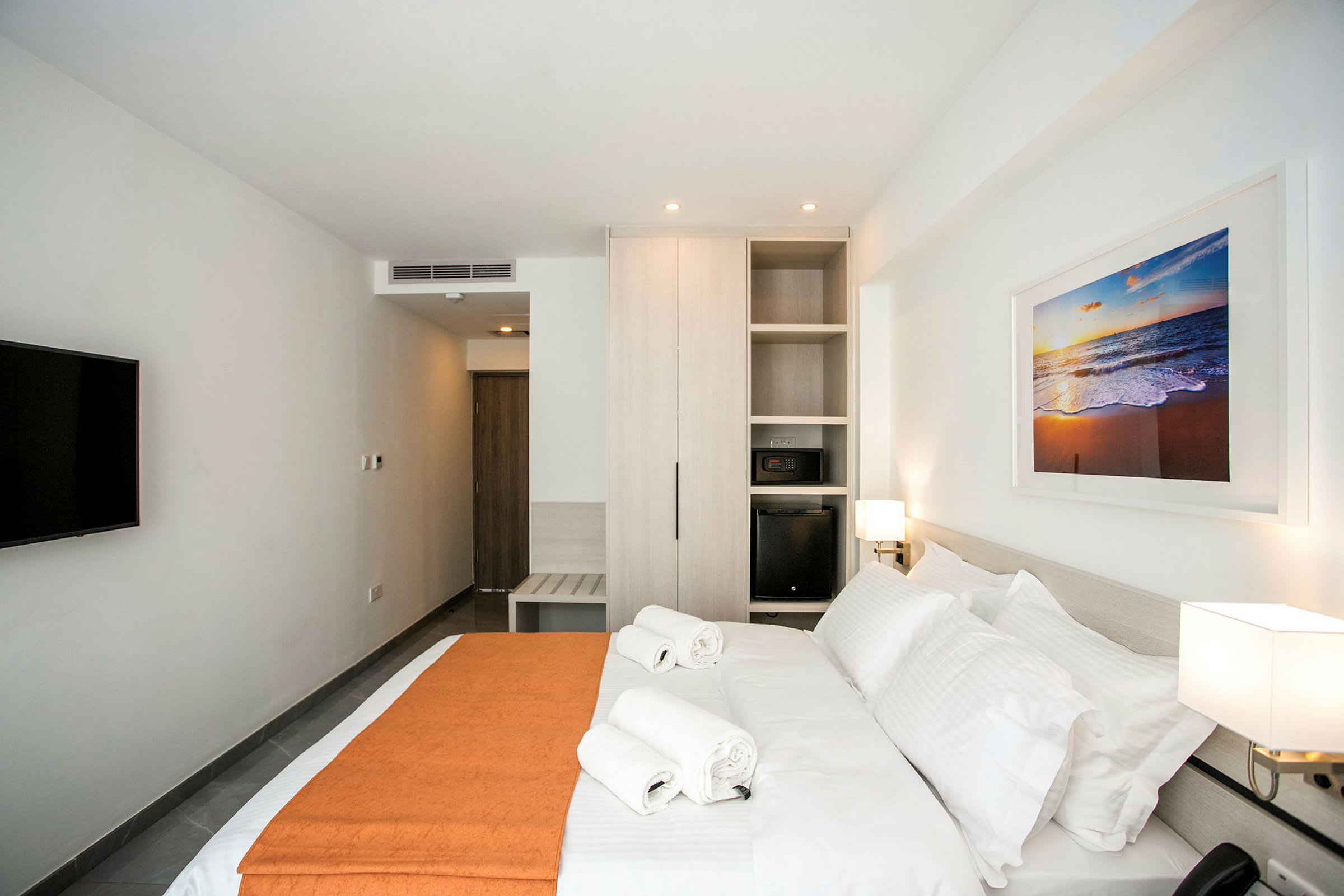 Ett modernt hotellrum med en stor säng, två handdukar ovanpå, en garderob, en TV på väggen och en tavla med motiv av en strand. (AI-genererad text)