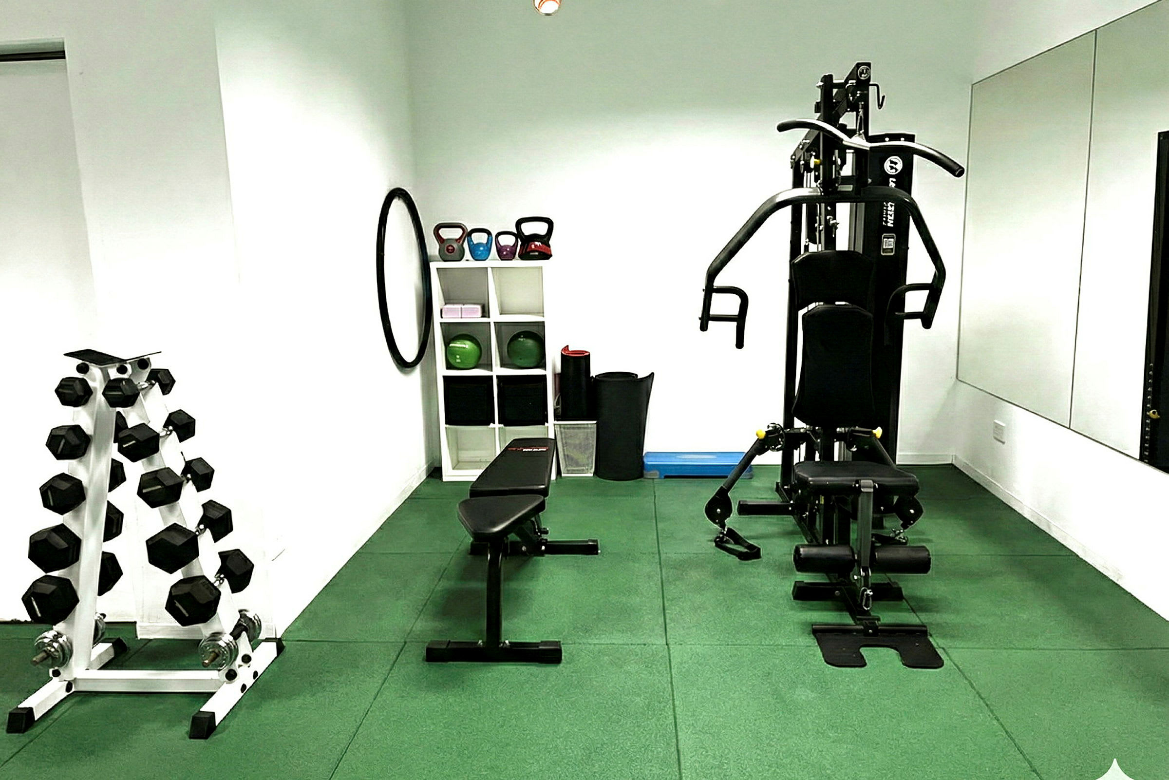 Ett gym med träningsutrustning som hantlar, bänk, kettlebells och en träningsmaskin. Rummet har gröna golvplattor och vita väggar. (AI-genererad text)