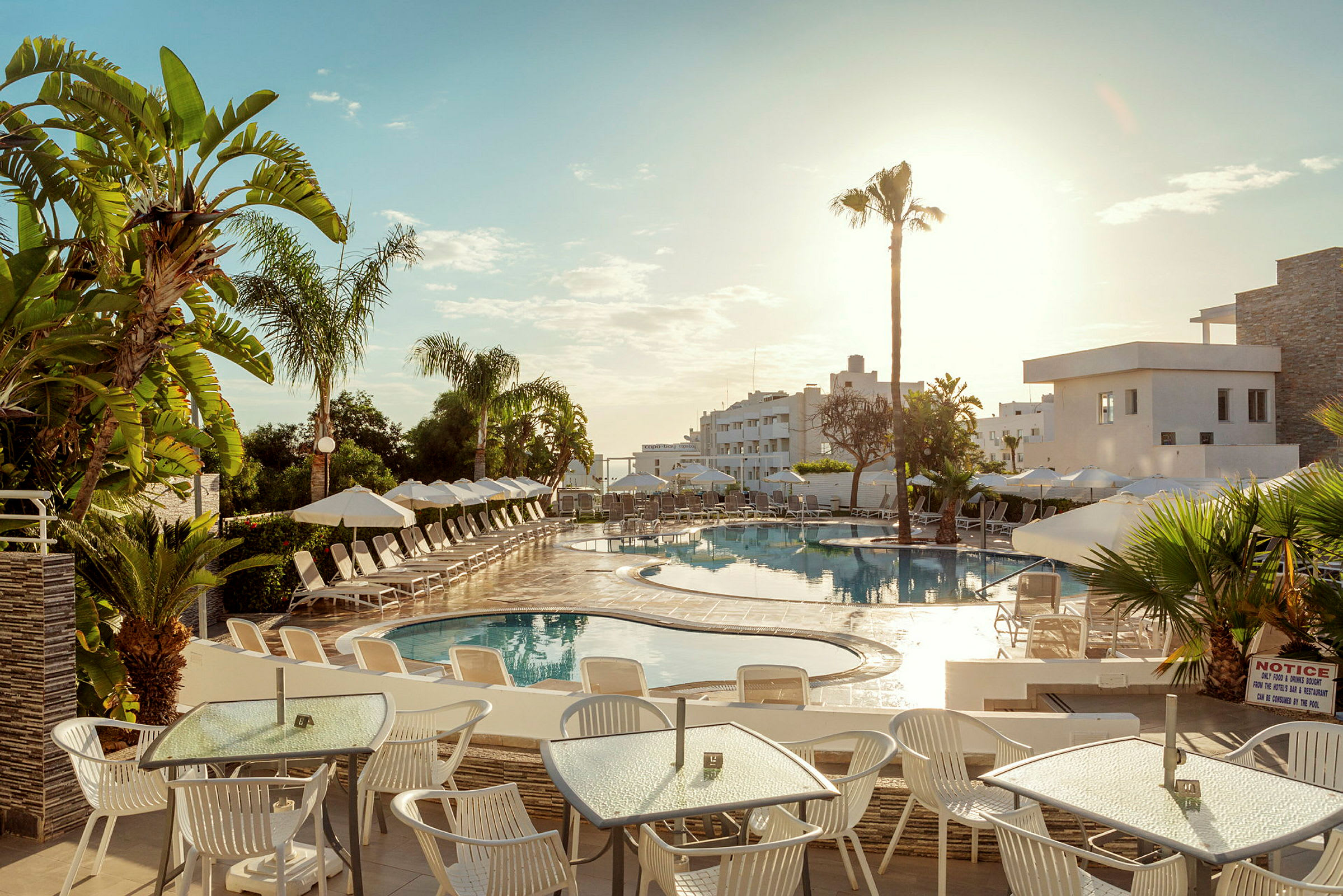 Bilden visar en utomhuspool på ett hotell, omgiven av solsängar och parasoller. I förgrunden finns bord och stolar under en solig himmel. (AI-genererad text)