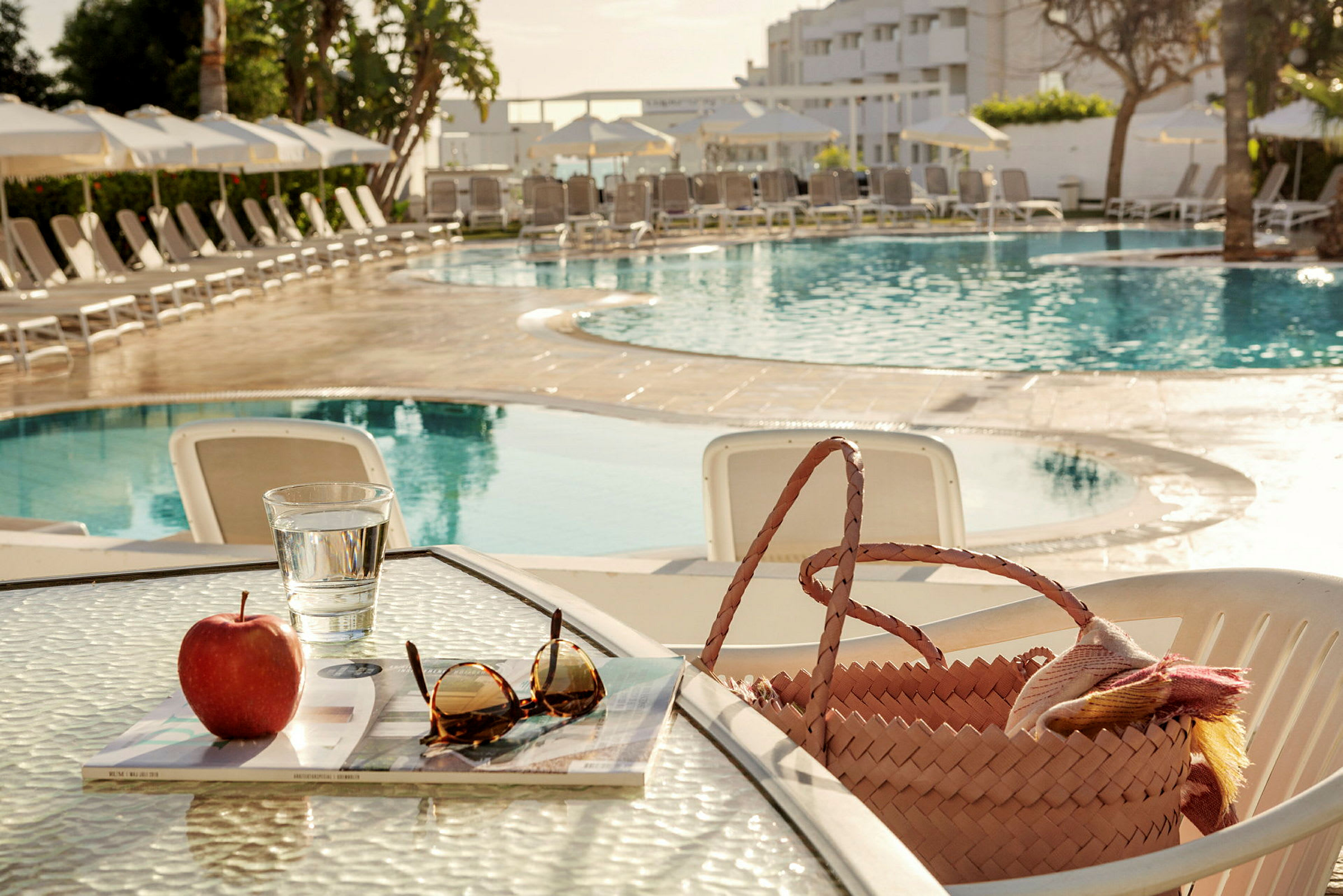 Ett bord med ett äpple, glas vatten, solglasögon och en korg som står bredvid en pool. (AI-genererad text)