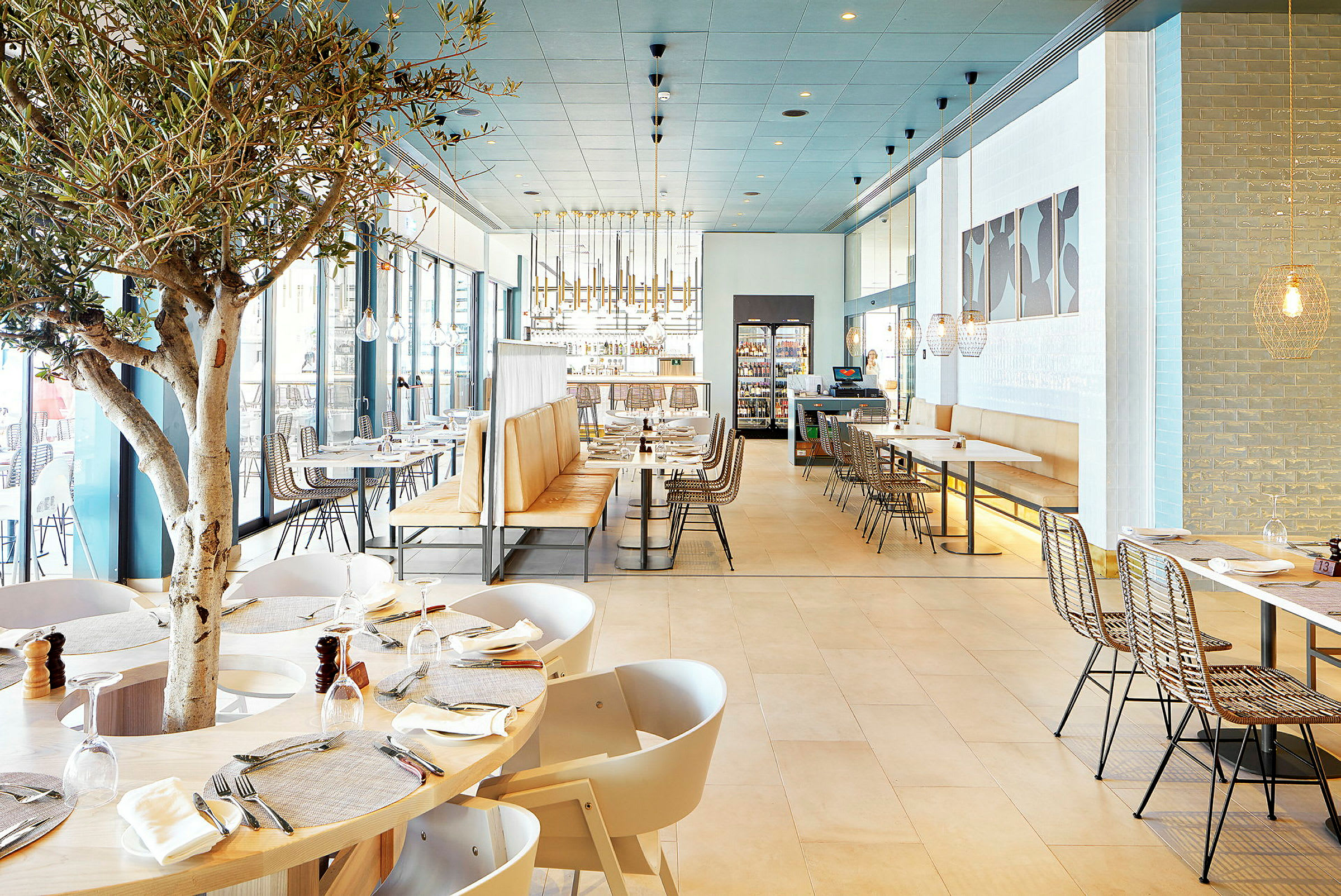 Interiör av en modern restaurang med trämöbler, ljusa färger och ett träd mitt bland borden. (AI-genererad text)