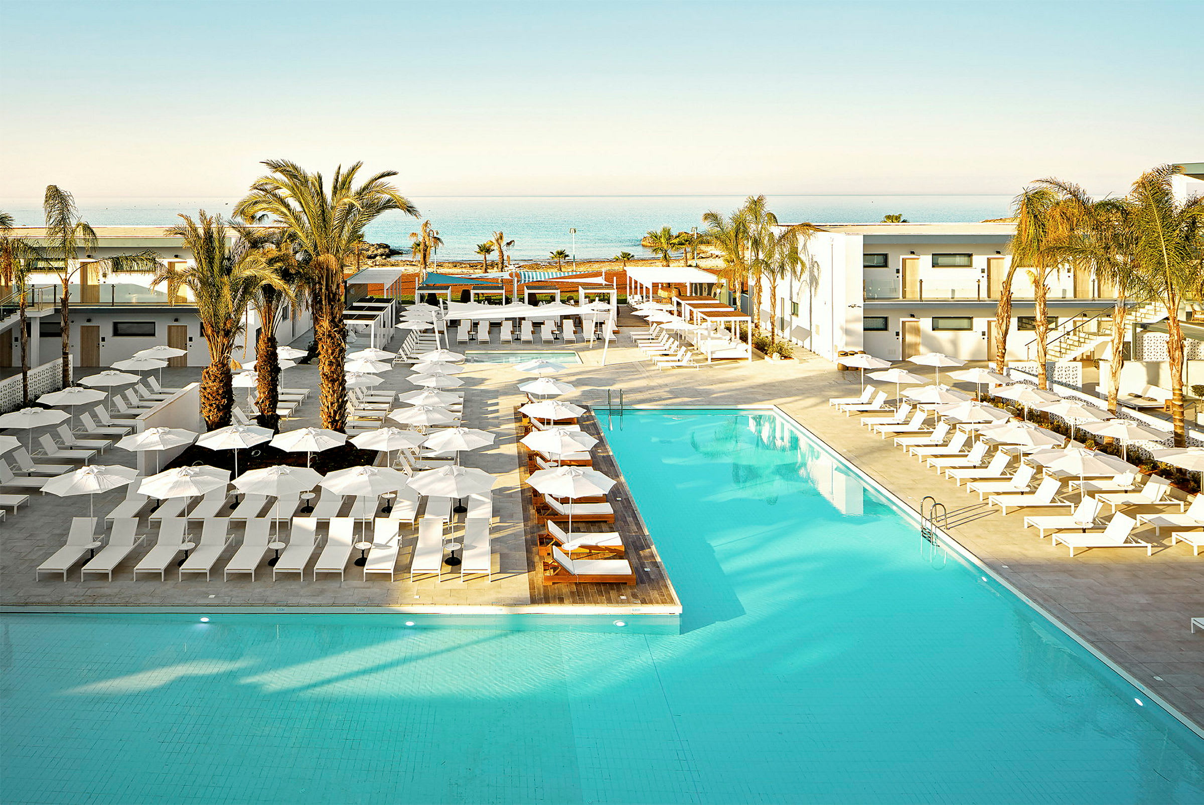 En hotellpool omgiven av solstolar och parasoller, med palmer och en havsutsikt i bakgrunden. (AI-genererad text)