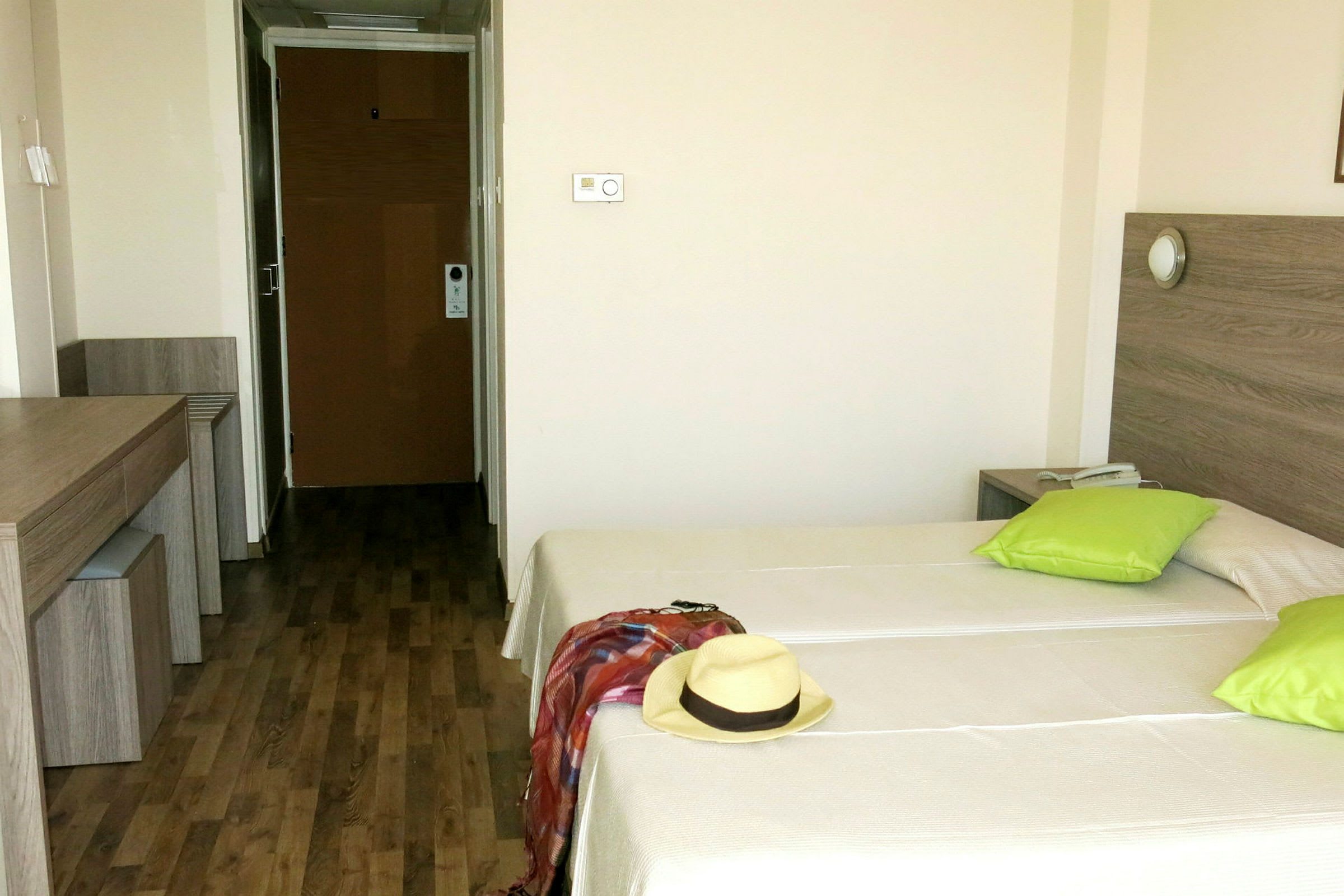 Ett hotellrum med två enkelsängar, gröna kuddar och en hatt och sjal på ena sängen. (AI-genererad text)