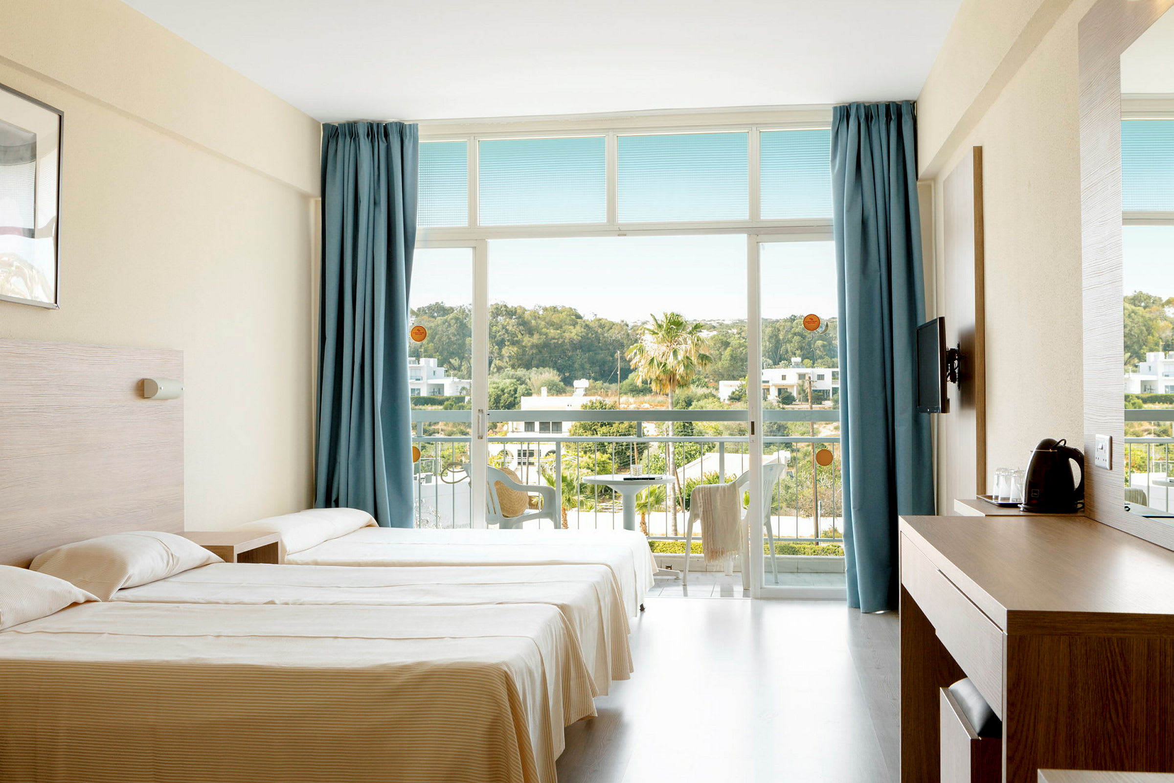 Ett hotellrum med två sängar, blå gardiner och en balkong med utsikt över grönska. (AI-genererad text)