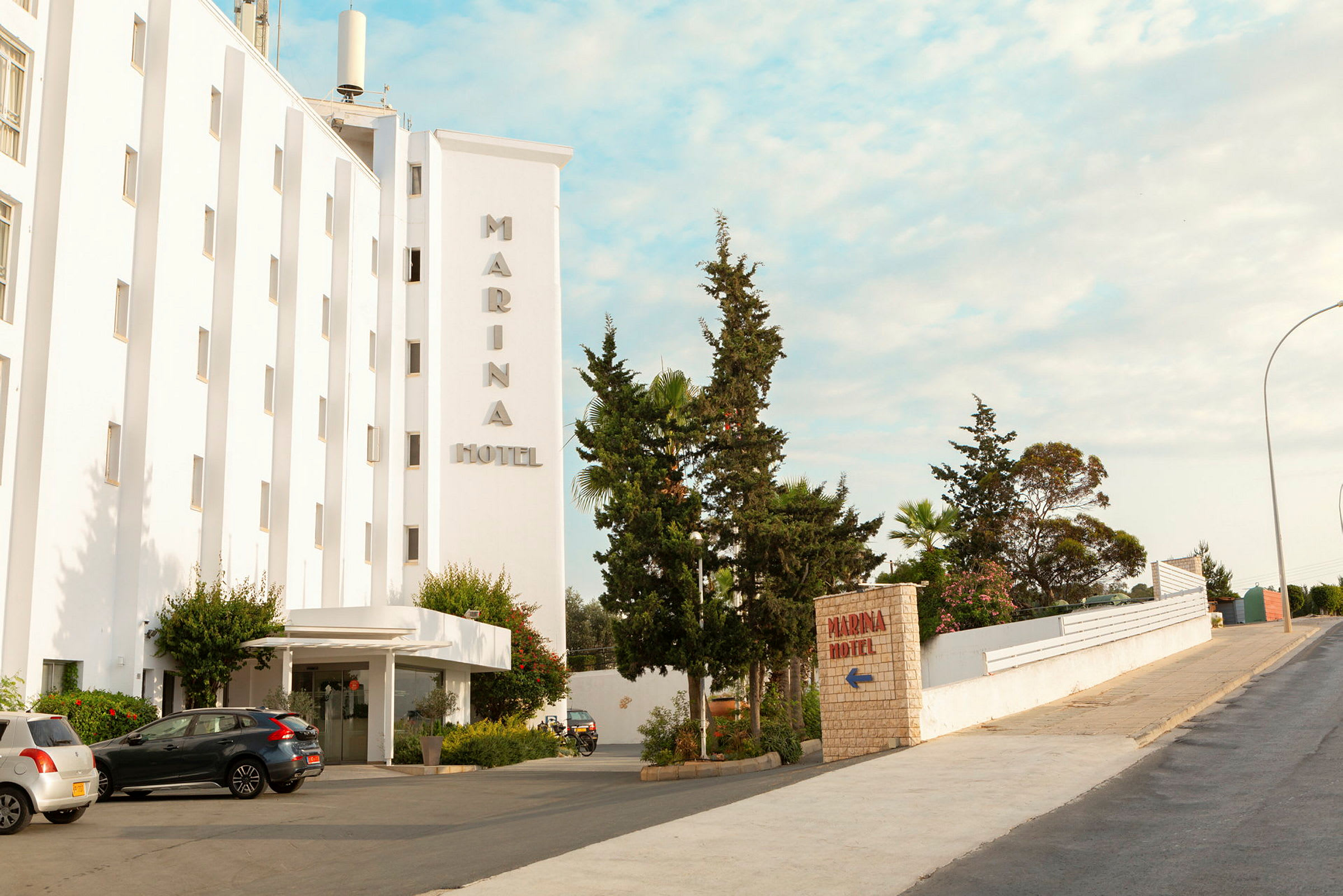 Utsikt över entrén till Marina Hotel med parkerade bilar och träd runt omkring. (AI-genererad text)