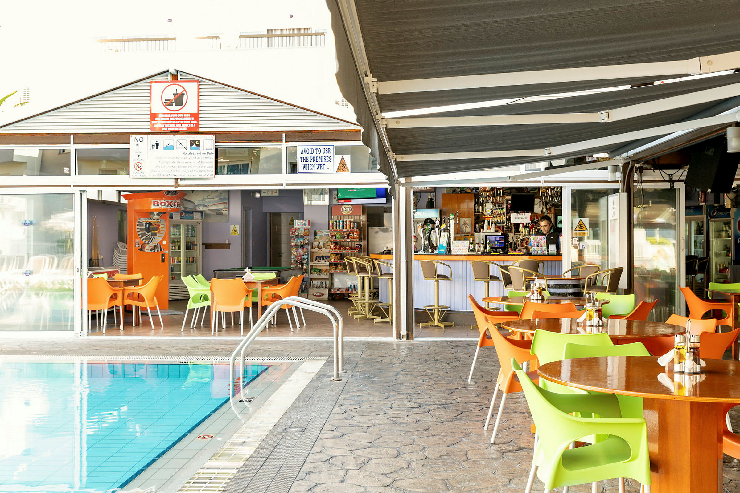 Utomhusområden med bord och stolar vid en pool, med en bar och matställe i bakgrunden. (AI-genererad text)