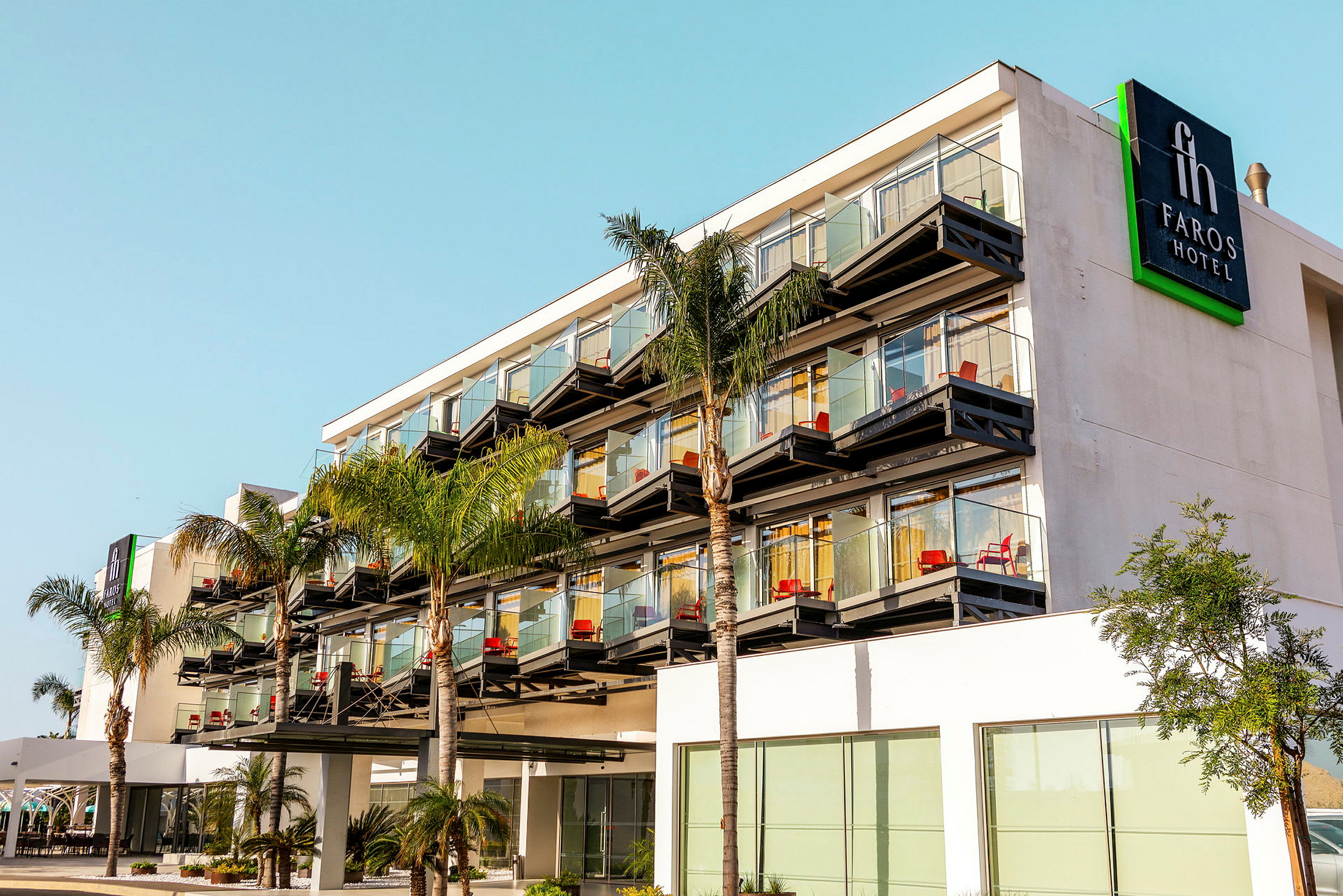 Utomhusbild av Faros Hotel med flera våningar, glasbalkonger och palmträd framför. (AI-genererad text)