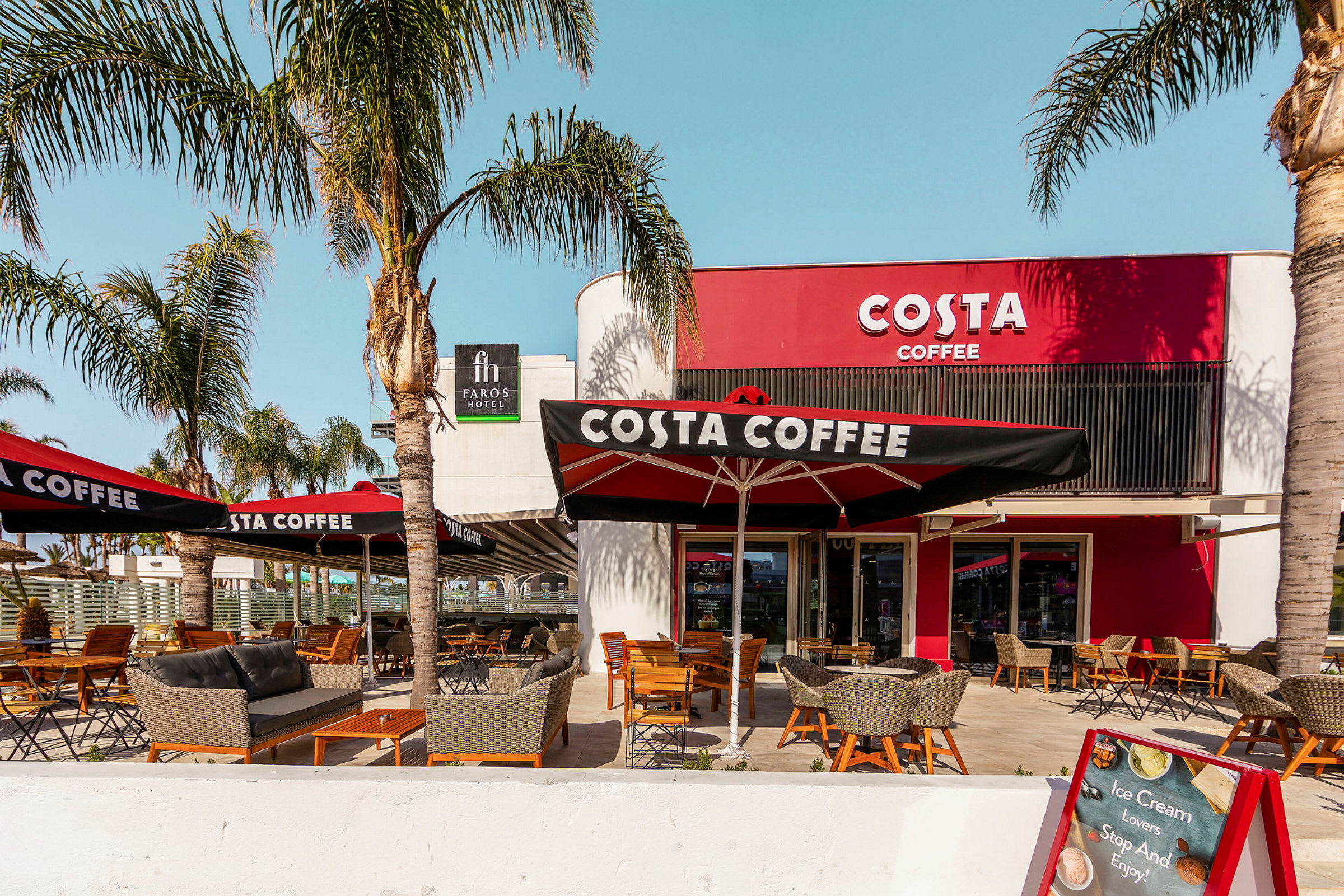 Utomhusservering vid kaféet Costa Coffee med palmer och sittmöbler. (AI-genererad text)