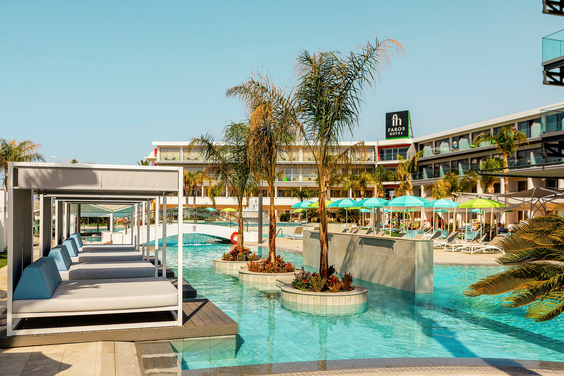 En pool omgiven av solstolar, parasoller och palmer framför en hotellbyggnad med balkonger. (AI-genererad text)