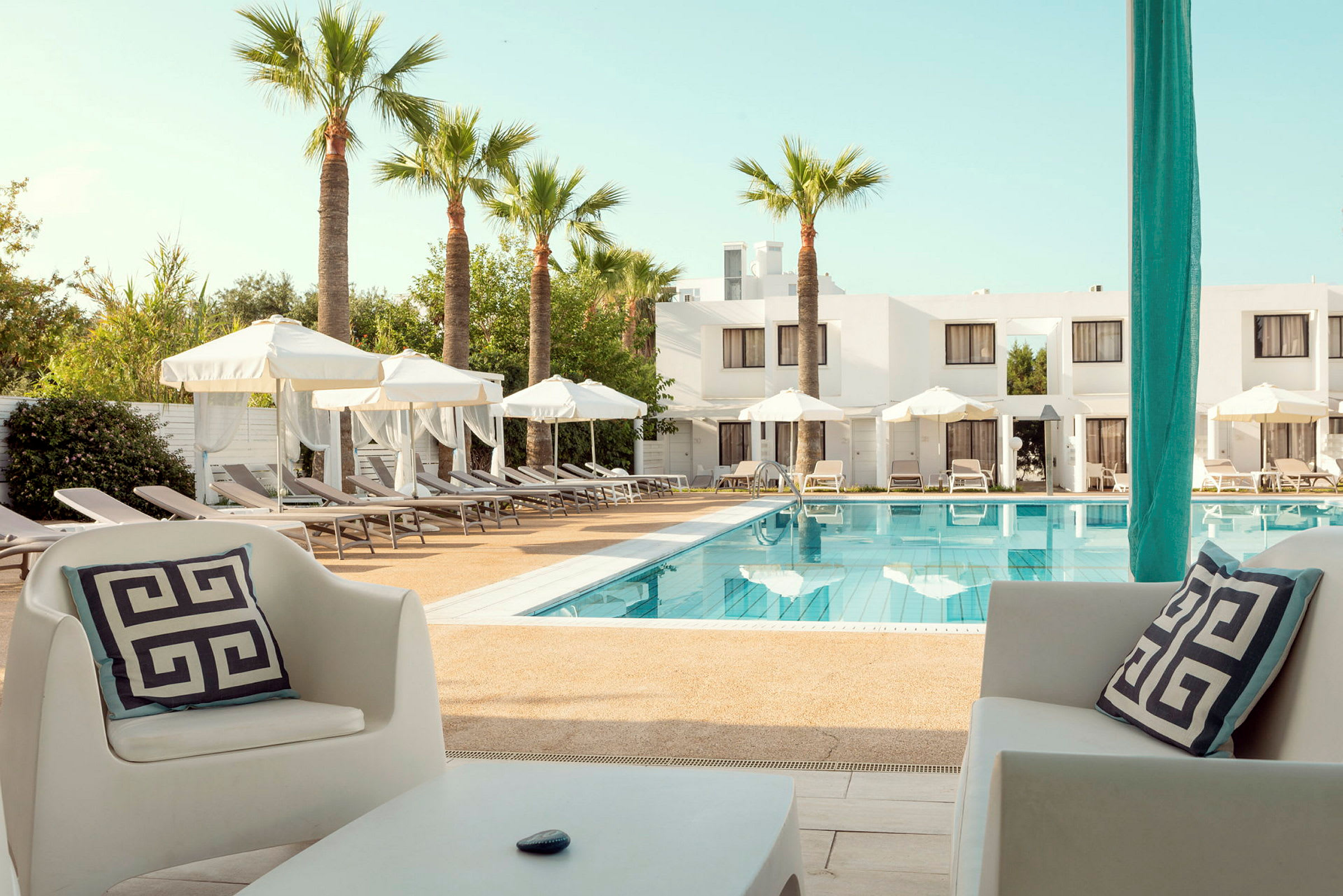 Vy över en hotellpool med solstolar och parasoller samt vita byggnader i bakgrunden. (AI-genererad text)