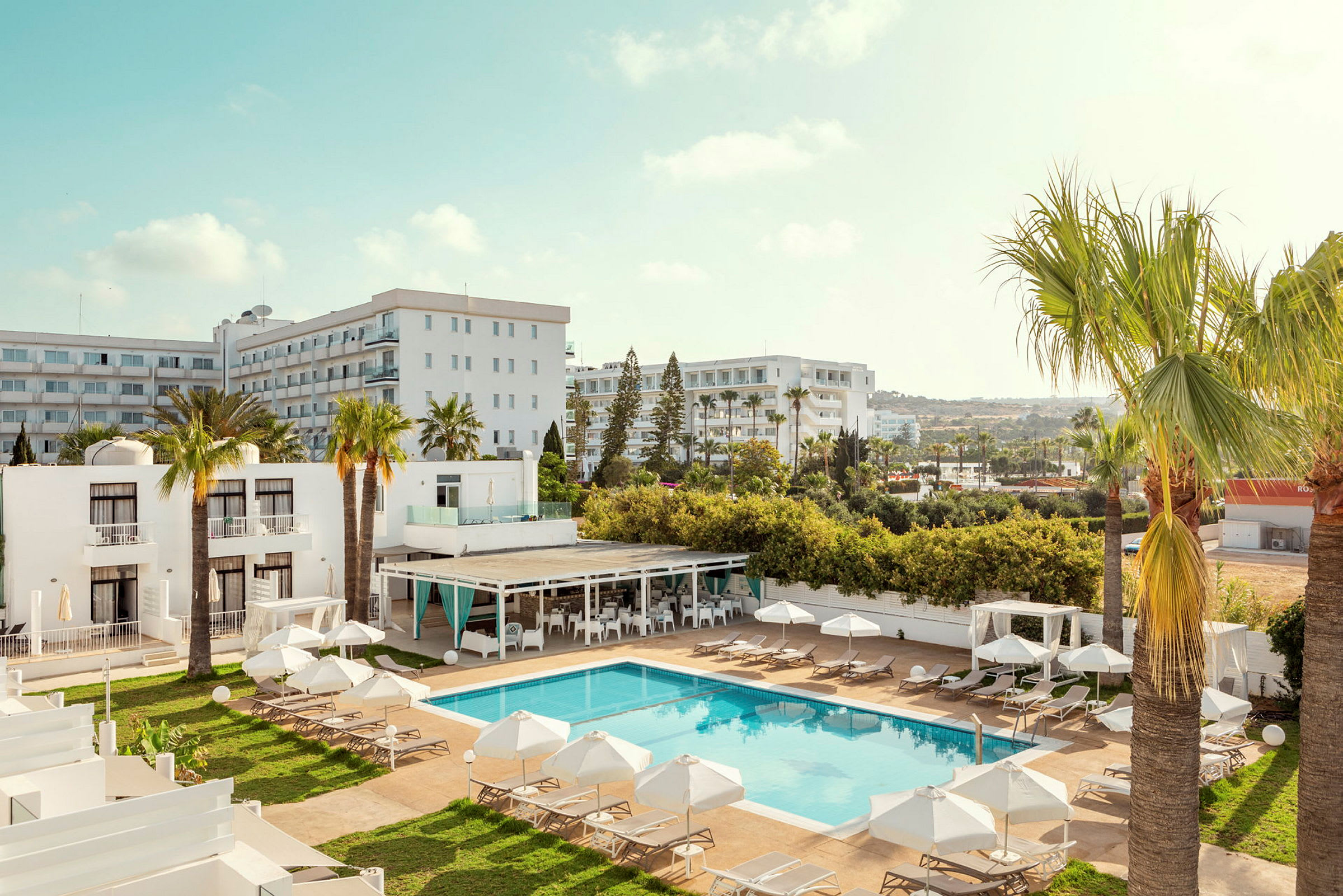 Ett hotellområde med pool, solstolar och parasoller, omgiven av palmer och byggnader. (AI-genererad text)