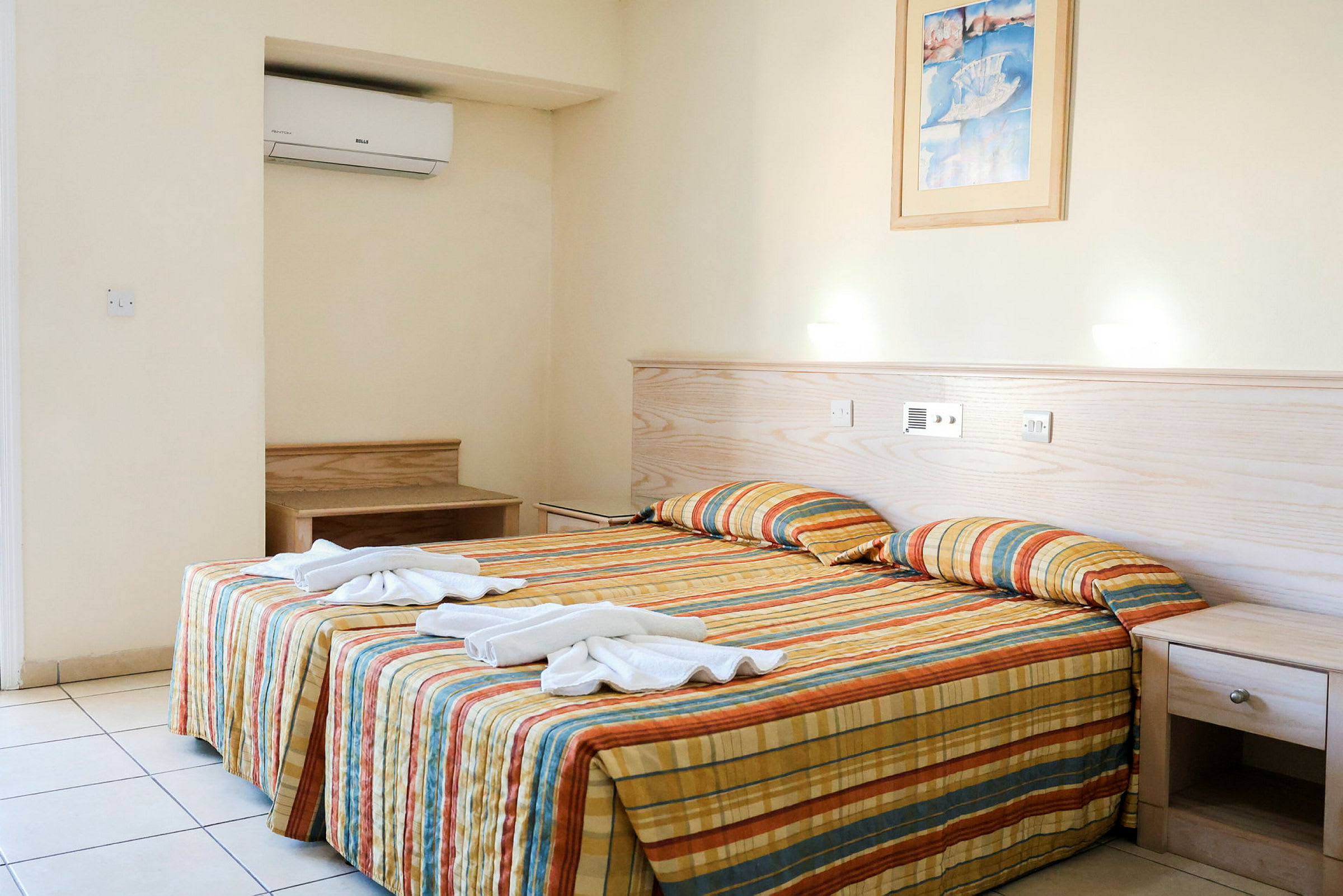 Ett hotellrum med en säng klädd i en färgglad randig överkast, två hopvikta handdukar placerade ovanpå, och en bild på väggen ovanför. Ett skrivbord och ett nattduksbord syns också. (AI-genererad text)