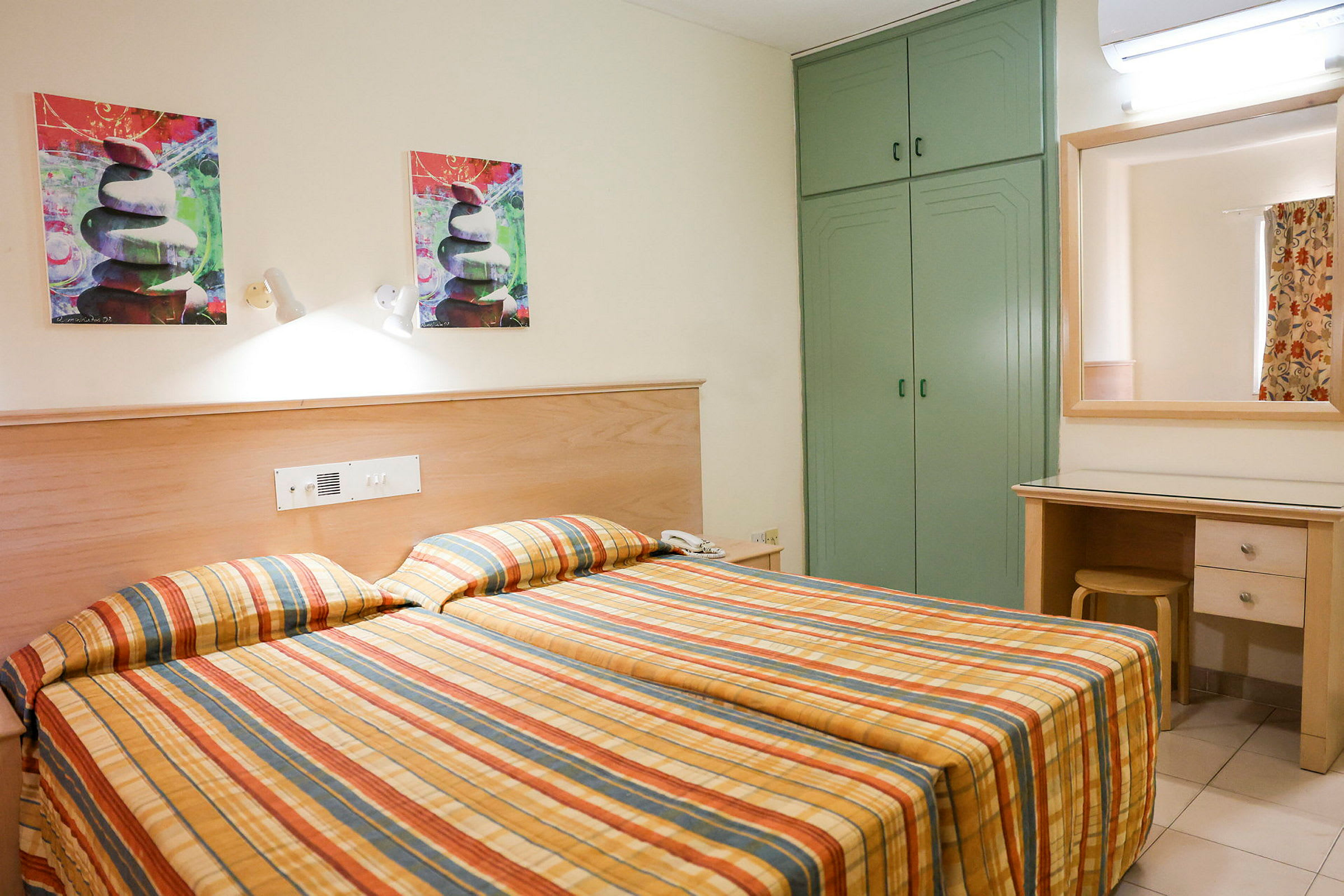 Hotellrum med två enkelsängar och rutiga sängöverdrag. Det finns en grön garderob, en spegel ovanför ett skrivbord och konstmålningar på väggen. (AI-genererad text)