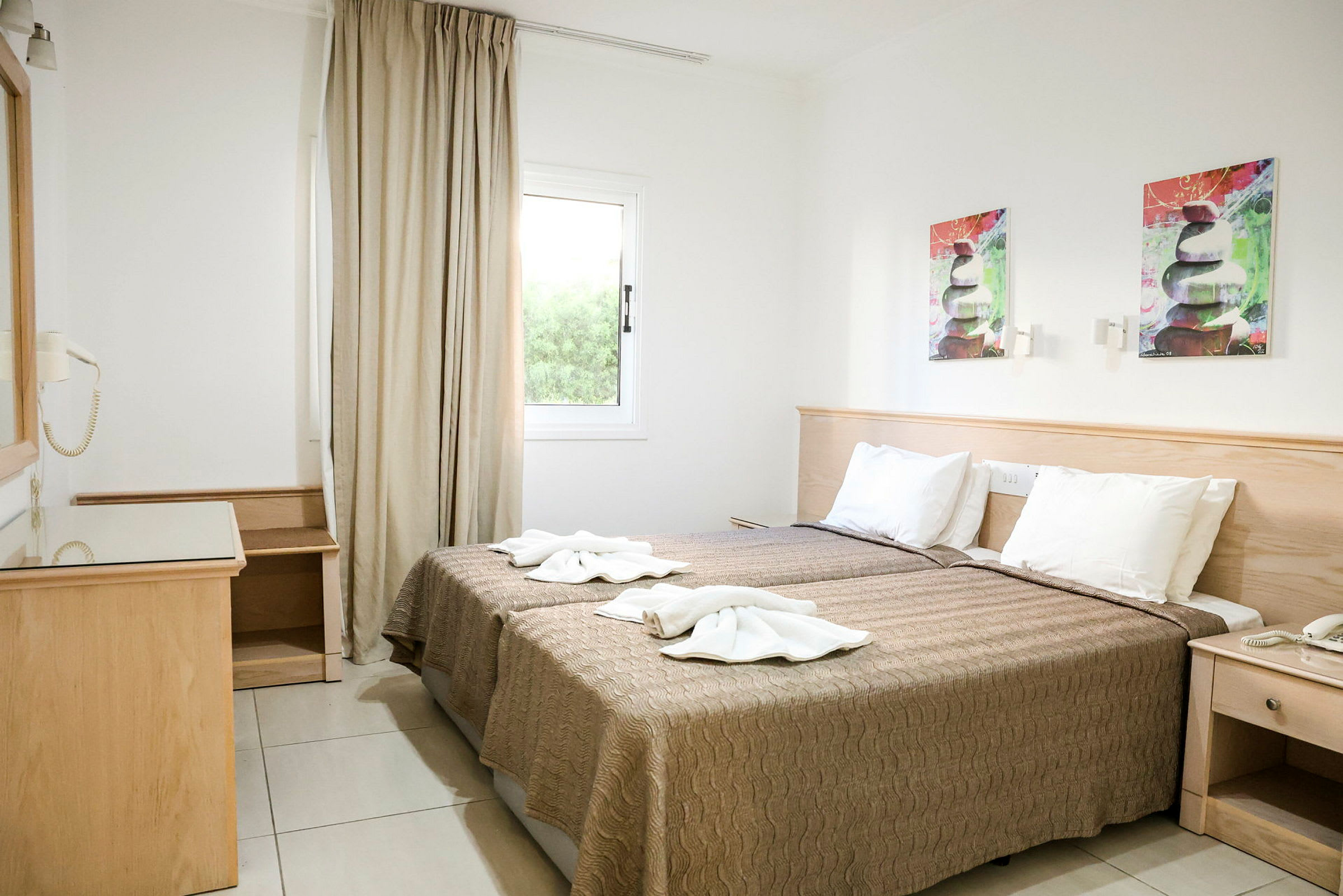 Bild av ett hotellrum med två enkelsängar. Rummet har vit vägg, ett fönster med beige gardiner och väggdekorationer ovanför sängen. Till vänster finns ett träskrivbord med spegel. (AI-genererad text)