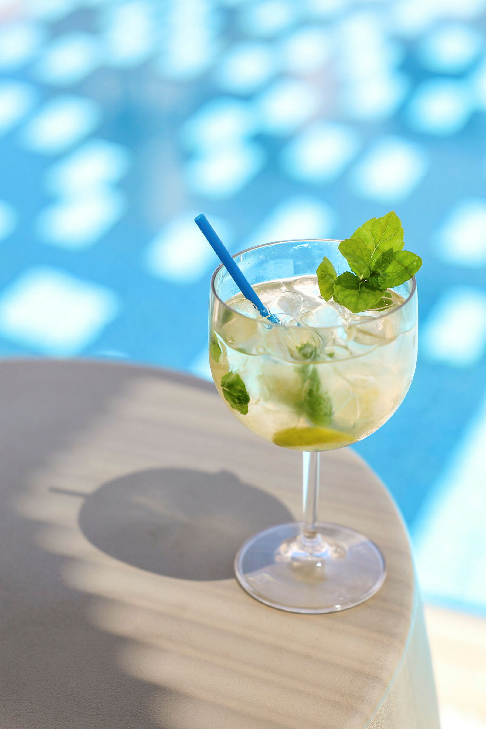 Ett glas med en kall dryck, dekorerad med en blå sugrör och färsk mynta, står på ett bord nära en swimmingpool. (AI-genererad text)