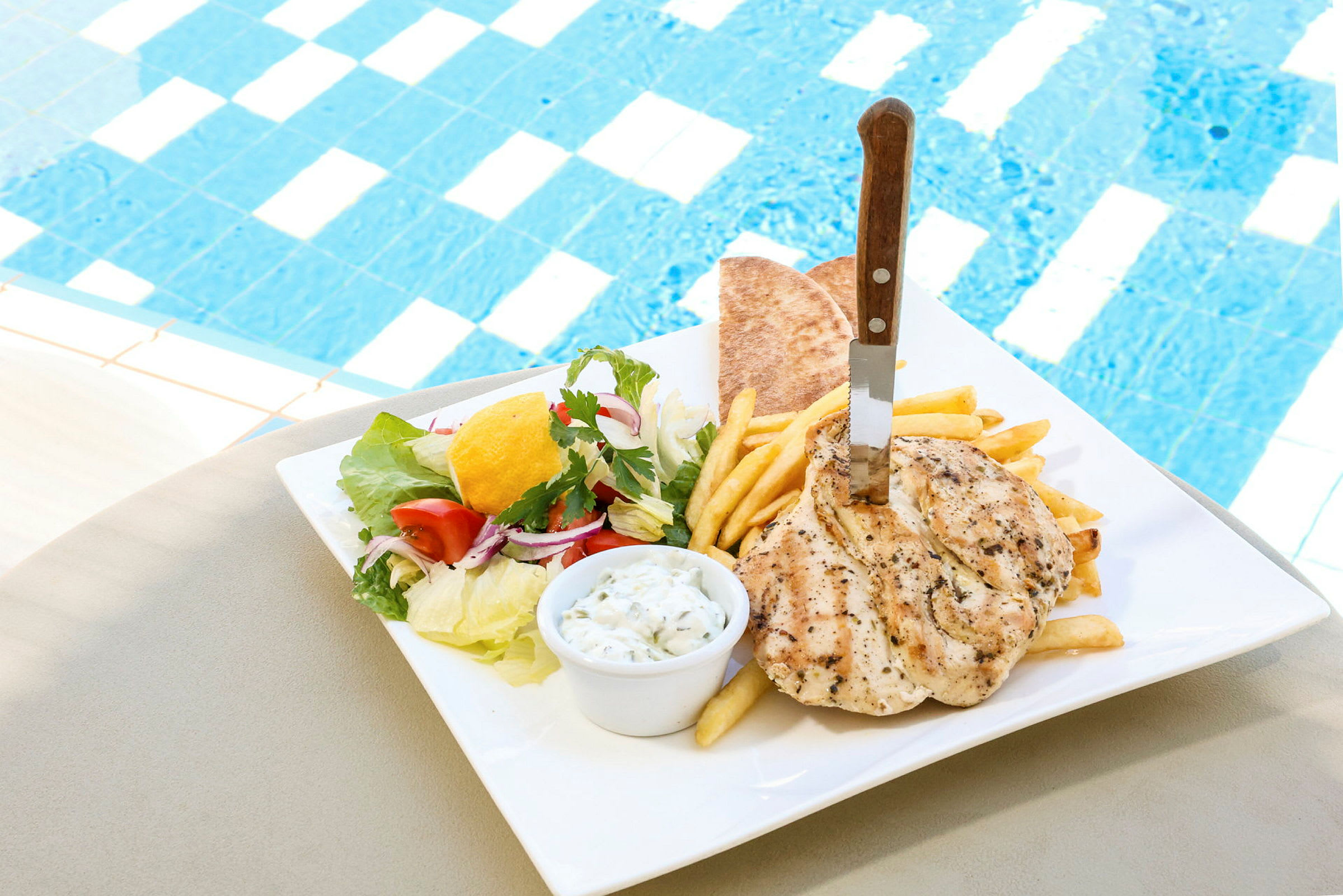 En tallrik med kyckling, pommes frites, pitabröd och sallad bredvid en pool. (AI-genererad text)