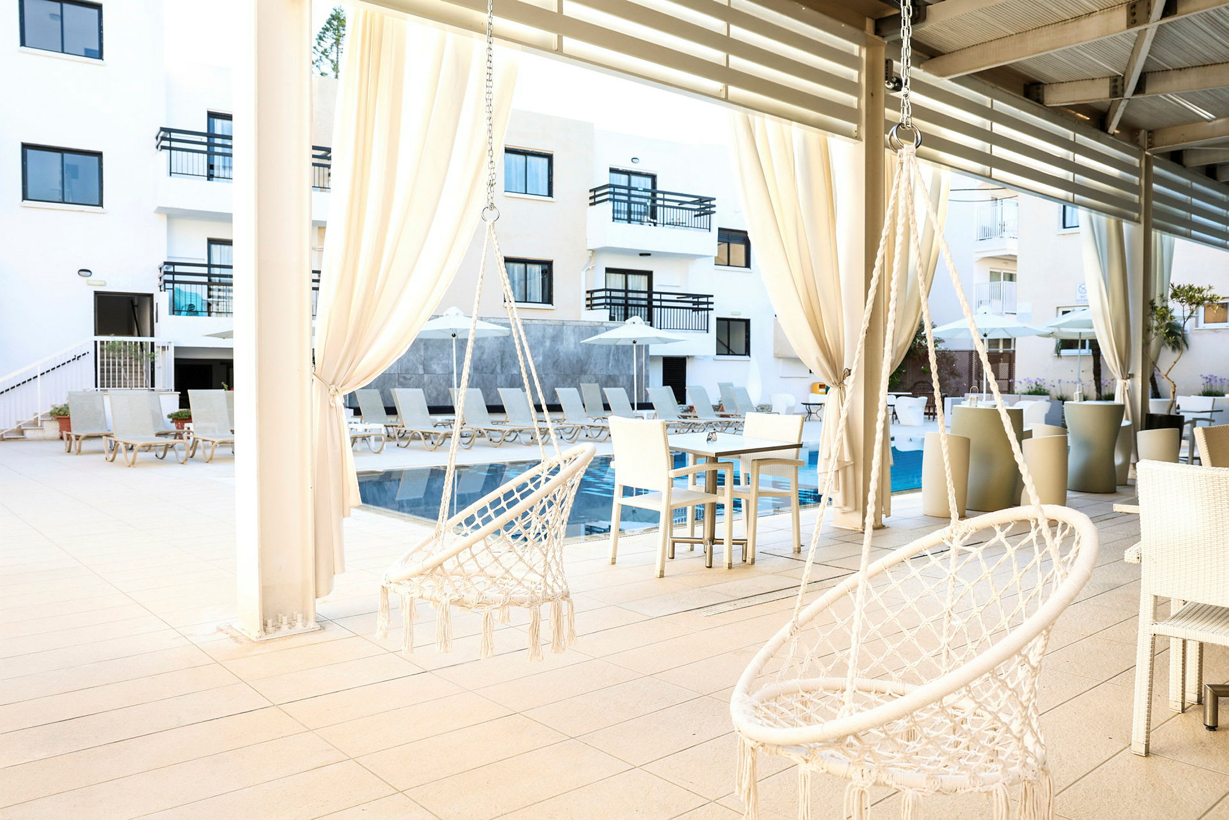 Bild av en solig hotellpool med solstolar och parasoller, samt två vita hammockstolar hängande under tak. (AI-genererad text)