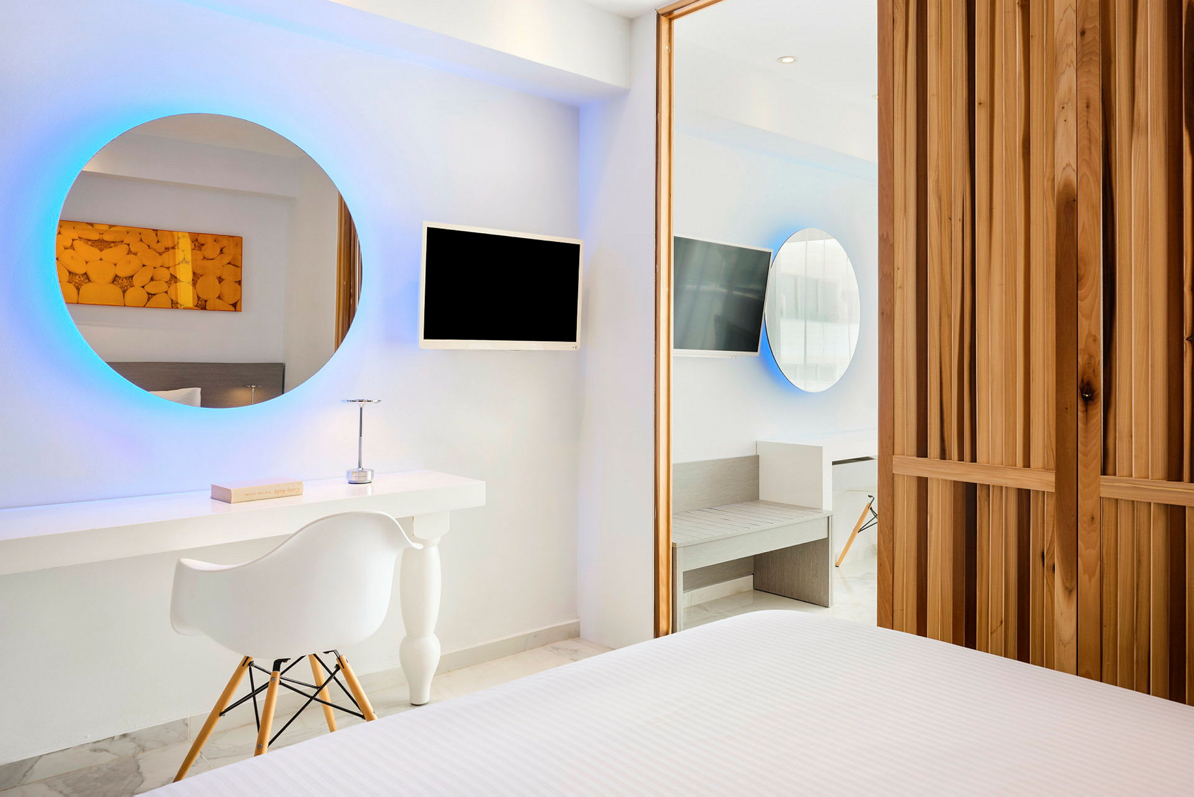 Ett modernt hotellrum med ett runt upplyst spegel, TV-skärm, ett skrivbord och en stol. Väggarna är vita och möblerna har minimalistisk stil. (AI-genererad text)