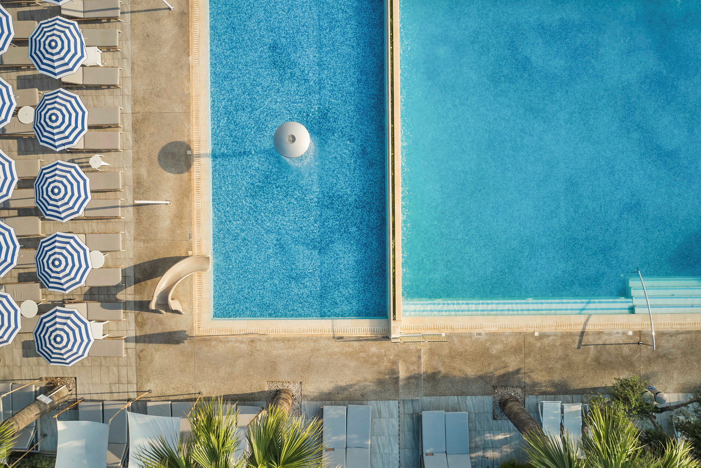 Flygfoto av en pool och solstolar med randiga parasoller. (AI-genererad text)