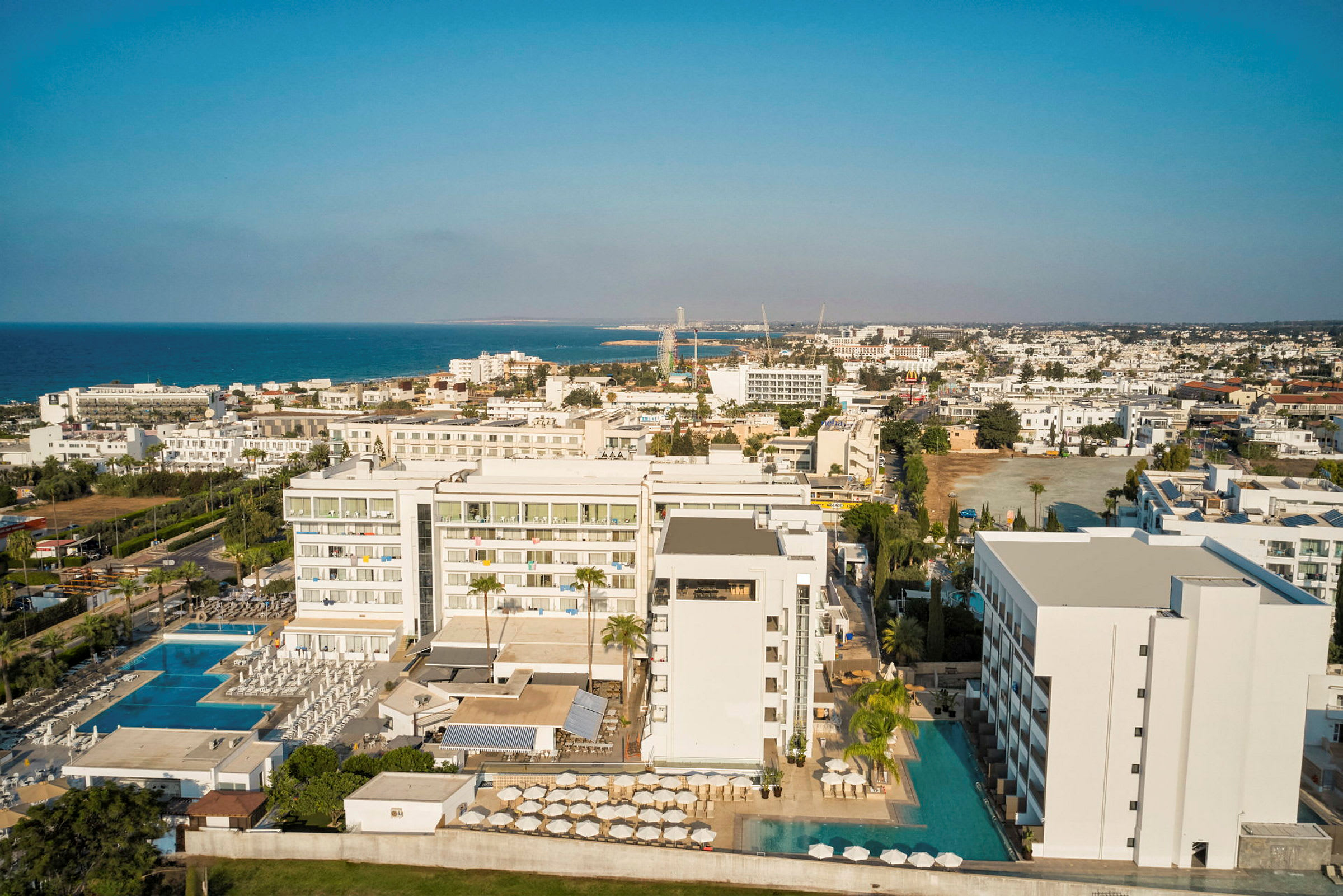 Flygbild av ett hotellkomplex nära havet med pooler och omgivande byggnader. (AI-genererad text)