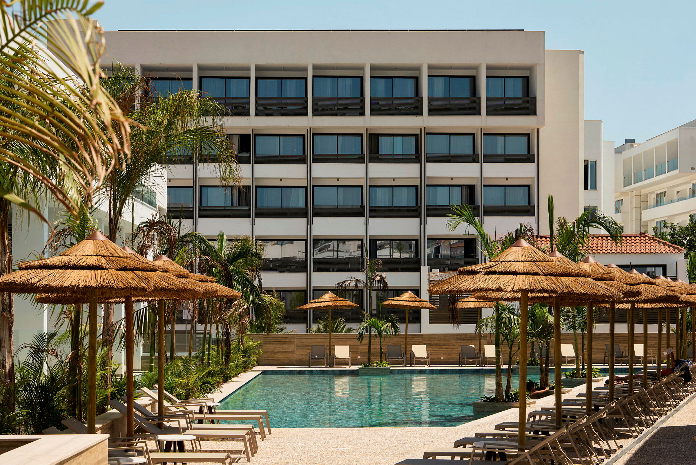 Ett hotell med en swimmingpool framför, omgiven av parasoll och solstolar. Det finns även palmer vid poolområdet. (AI-genererad text)