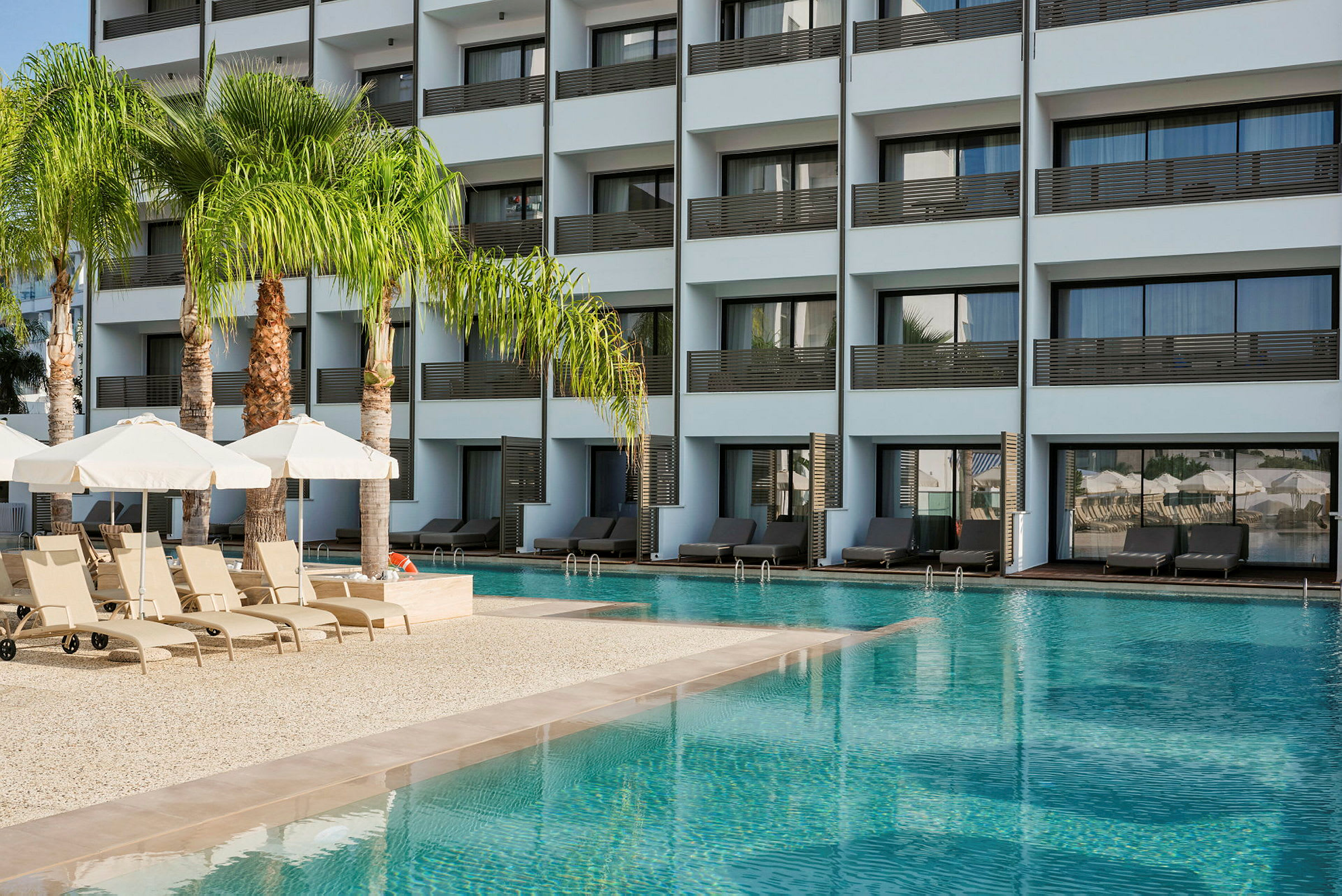 En pool med solstolar och parasoller framför en modern byggnad med balkonger. (AI-genererad text)
