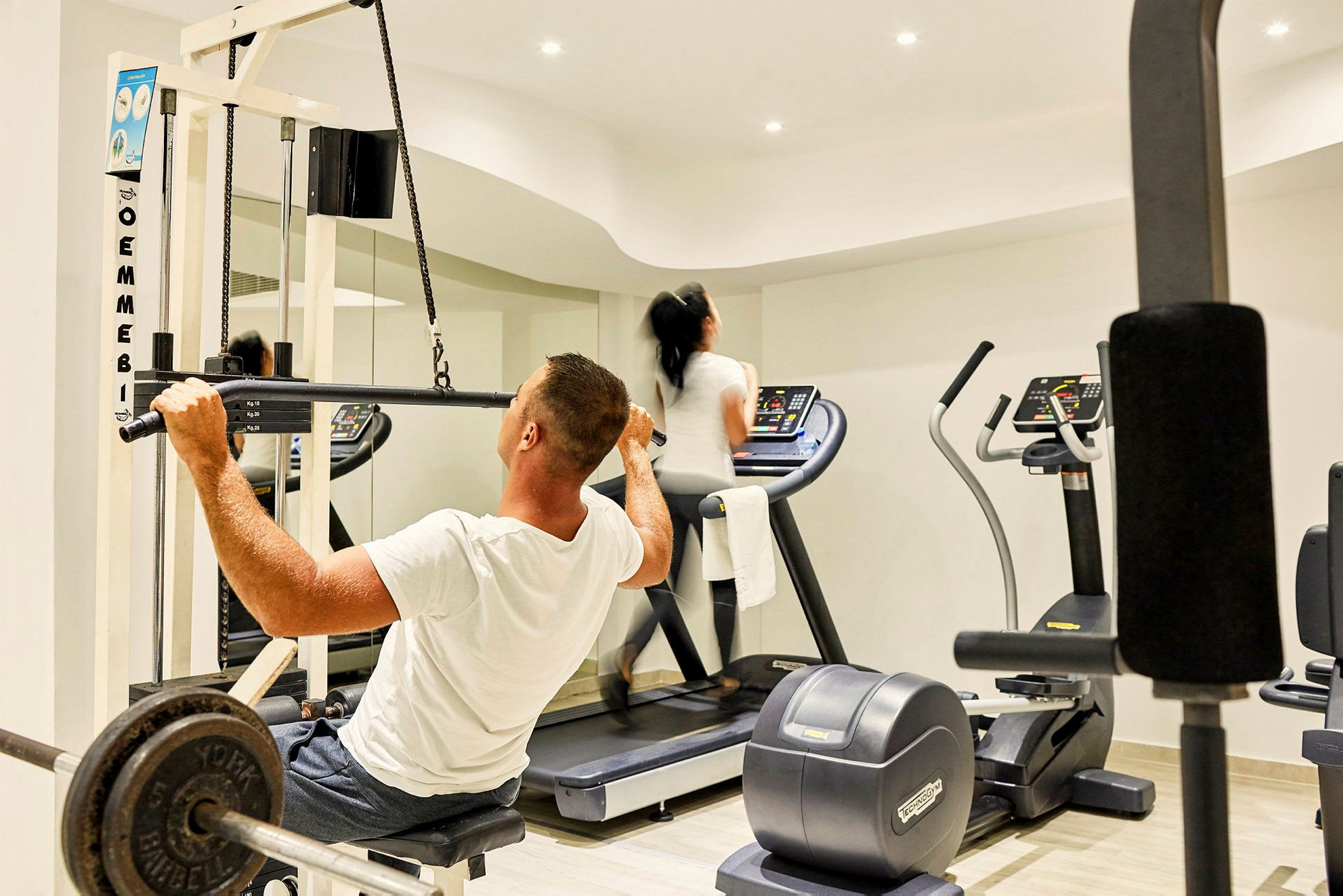 Ett gym med två personer som tränar, en på ett löpband och den andra som använder en dragmaskin. (AI-genererad text)