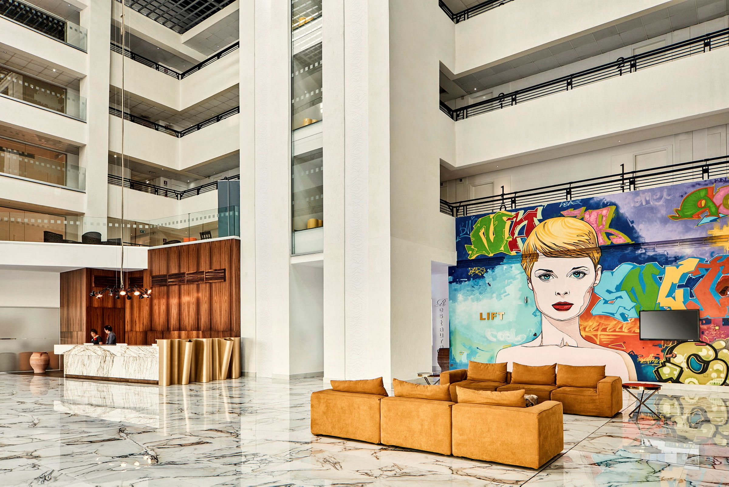 Interiör av ett modernt hotellatrium med marmorgolv, en sittgrupp med orange soffor och en stor väggmålning med en person. (AI-genererad text)
