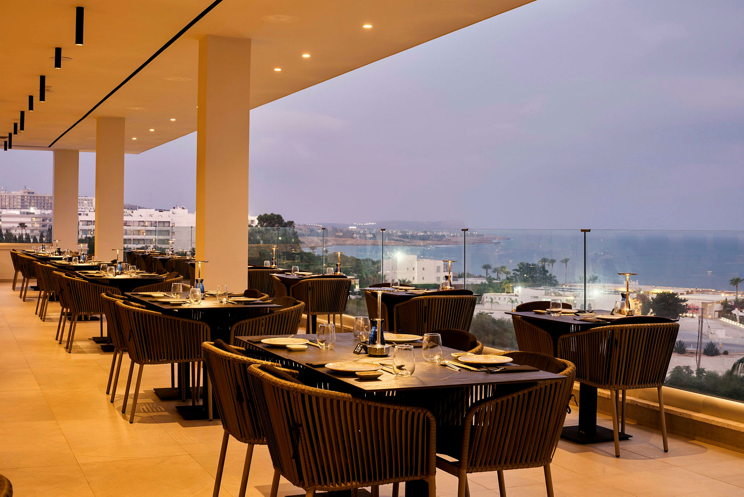 En restaurang med uteservering har bord och stolar uppradade, med utsikt över havet och staden. (AI-genererad text)
