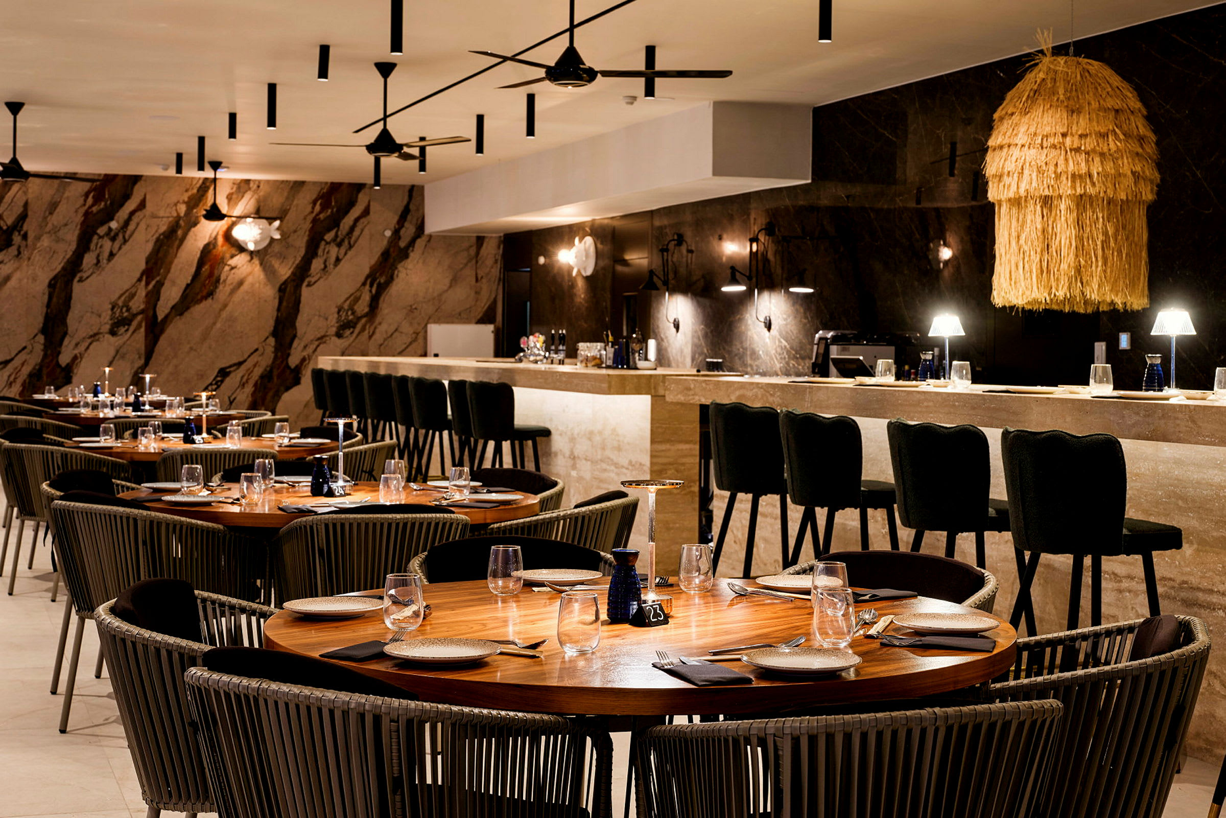 Ett elegant restaurangrum med brickbord och stolar, en bar i bakgrunden och stämningsfull belysning med en halminspirerad lampa. (AI-genererad text)