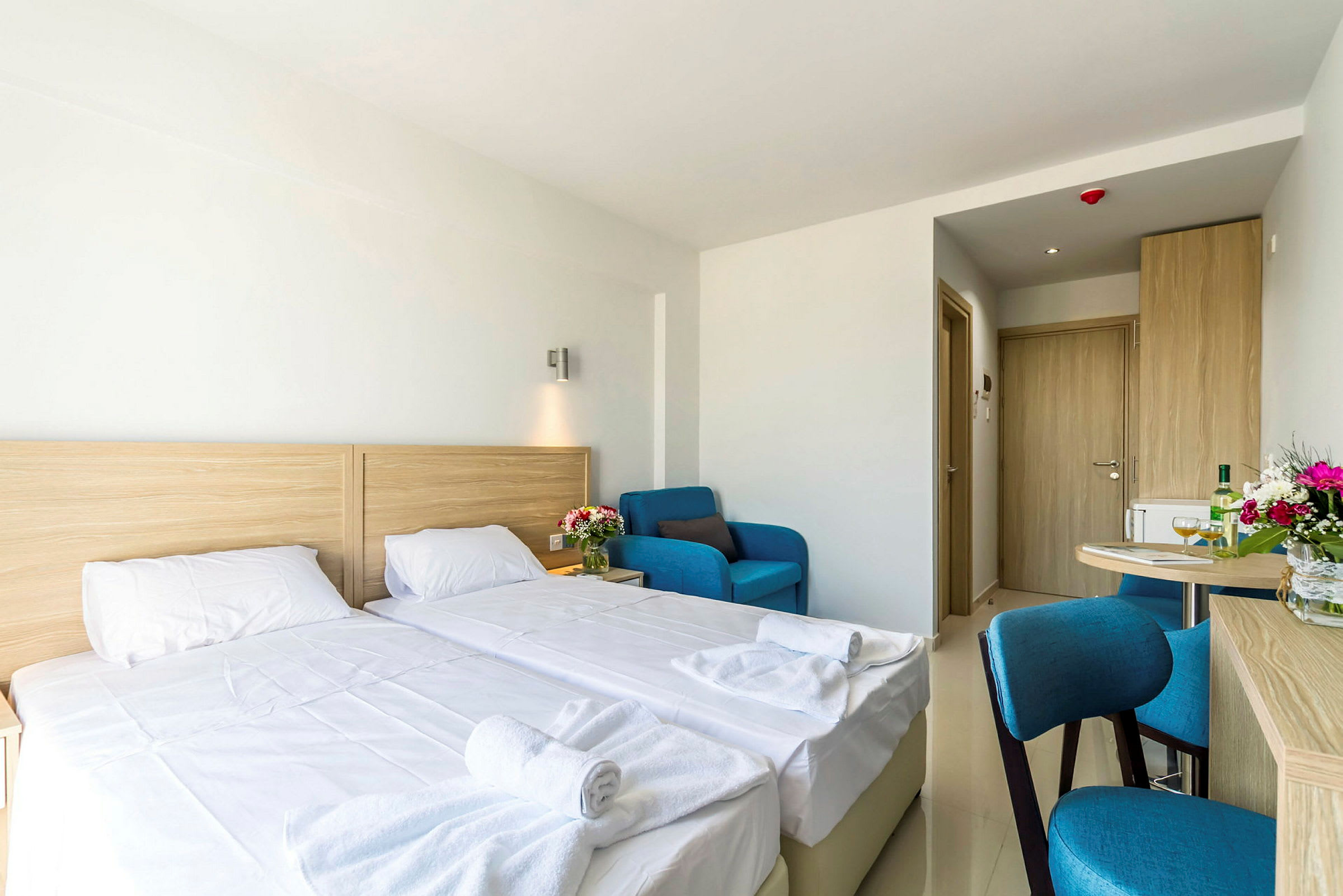 Ett hotellrum med två enkelsängar, en blå fåtölj, ett bord och en dörr i bakgrunden. (AI-genererad text)
