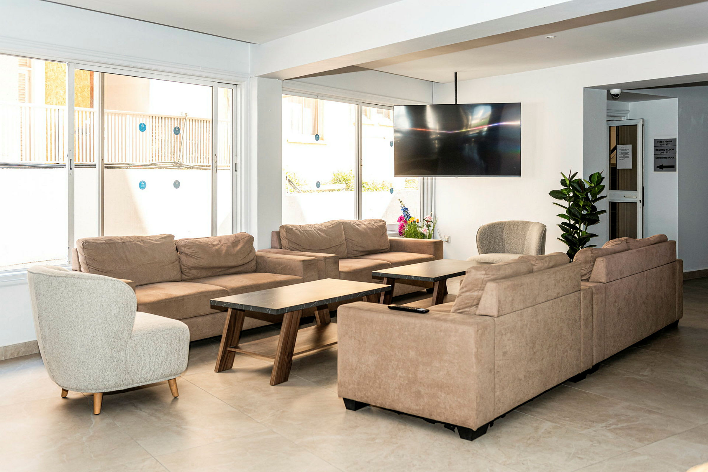 Ett vardagsrum med beige soffor, ett runt fåtölj, soffbord och en stor plattskärms-TV på väggen samt växter som dekoration. (AI-genererad text)
