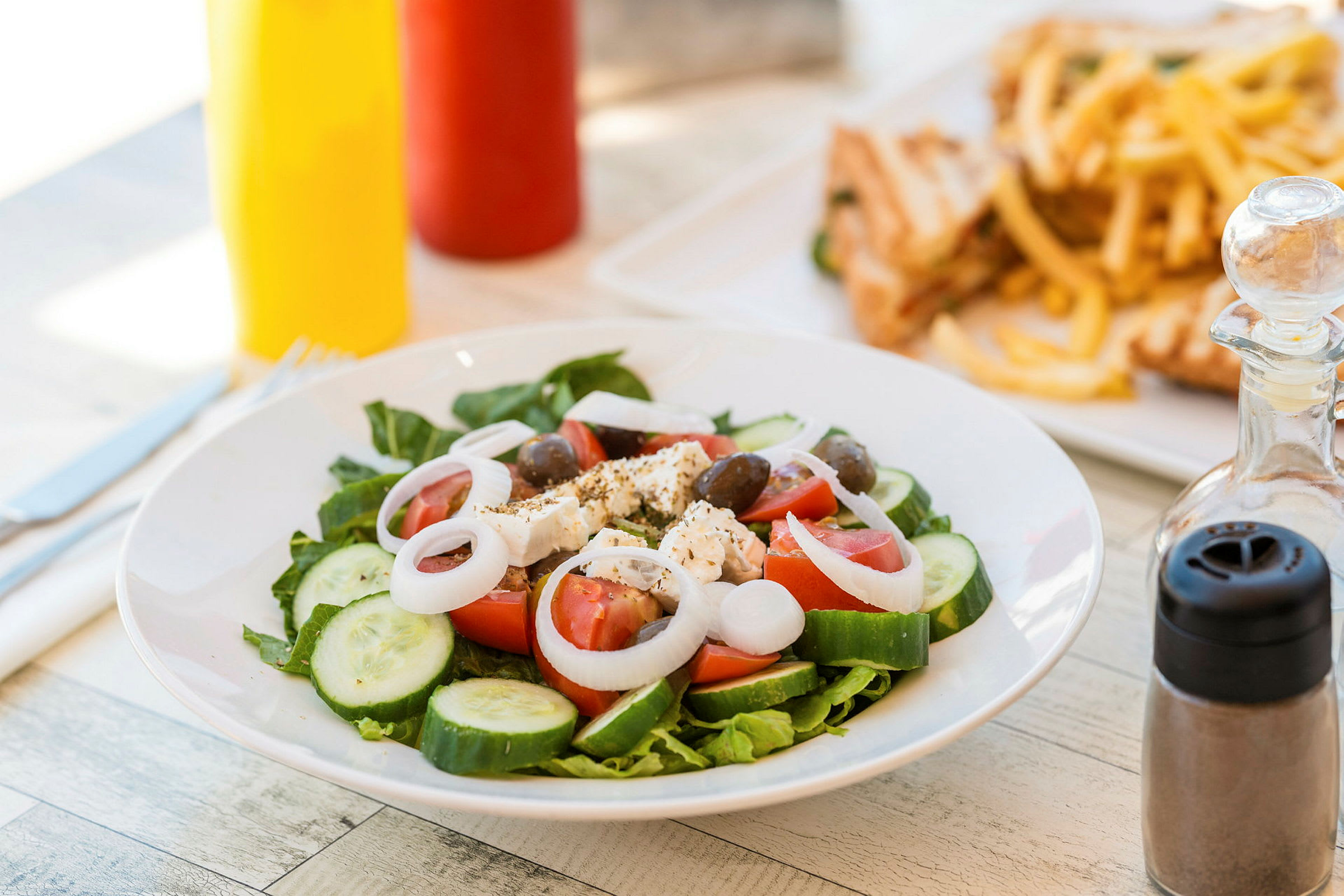 En tallrik med sallad innehållande tomat, gurka, lök, fetaost och oliver. I bakgrunden syns pommes frites och smörgåsar. (AI-genererad text)