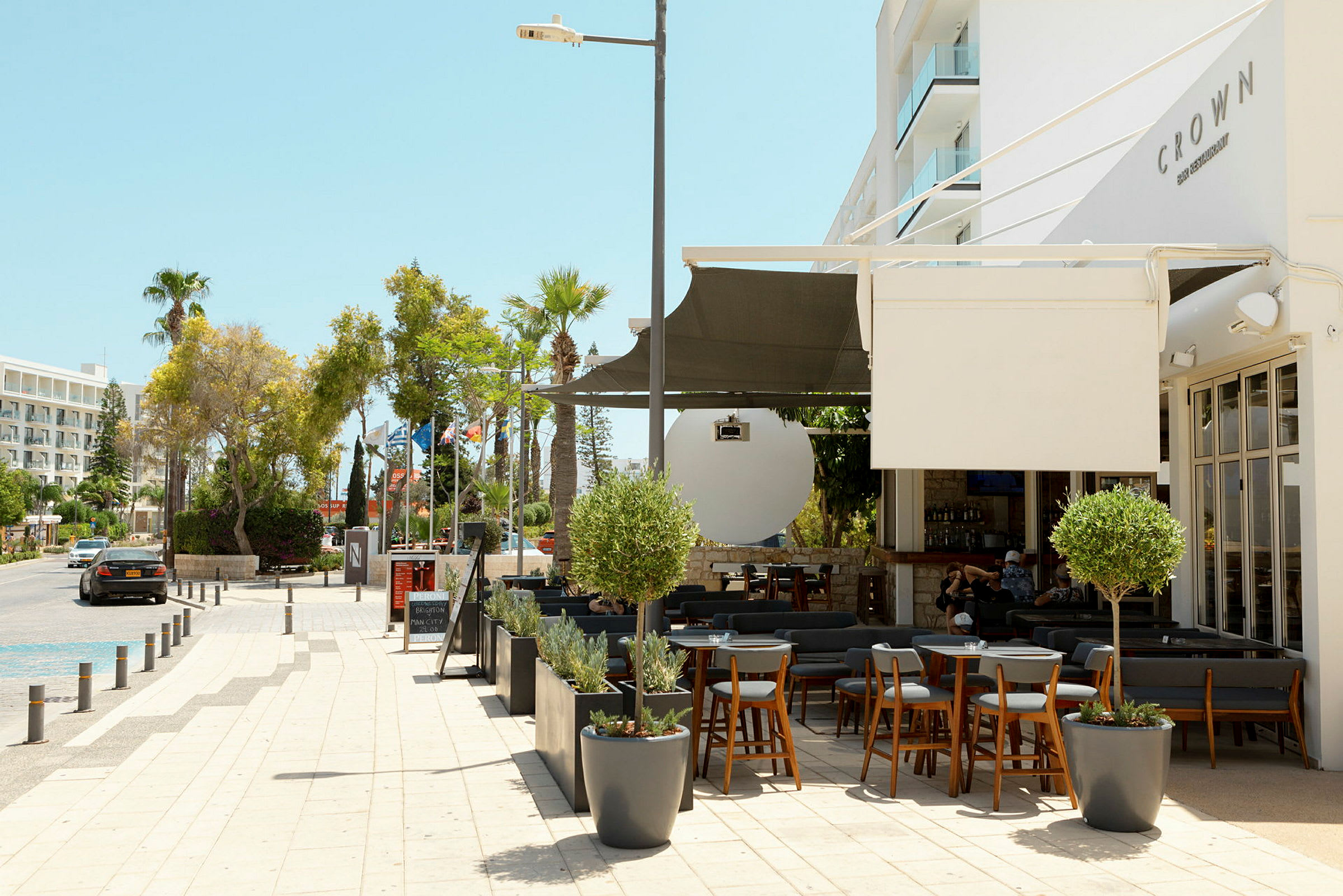 Bild av en utomhusrestaurang och trottoar intill en gata med träd och byggnader i bakgrunden. (AI-genererad text)