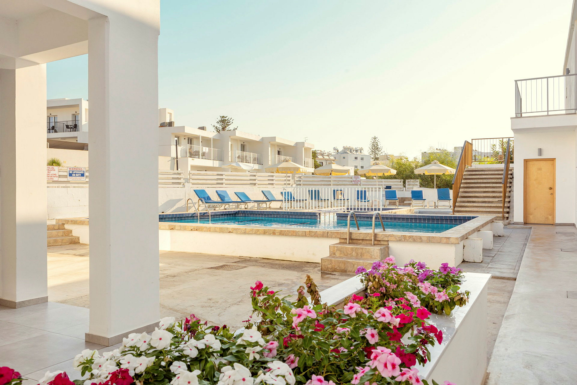 Bild av en hotellpool med solstolar och parasoller, omgiven av vita byggnader och blommor i förgrunden. (AI-genererad text)