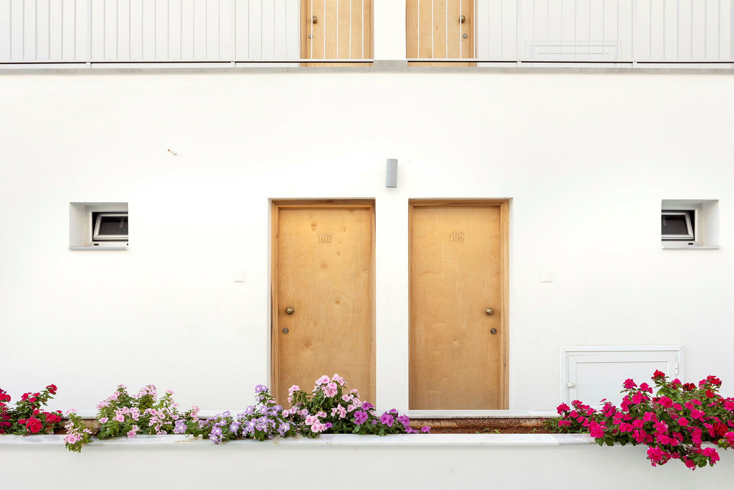 Två trädörrar med nummer 107 och 108 på en vit vägg, omgivet av flerfärgade blommor utanför. (AI-genererad text)