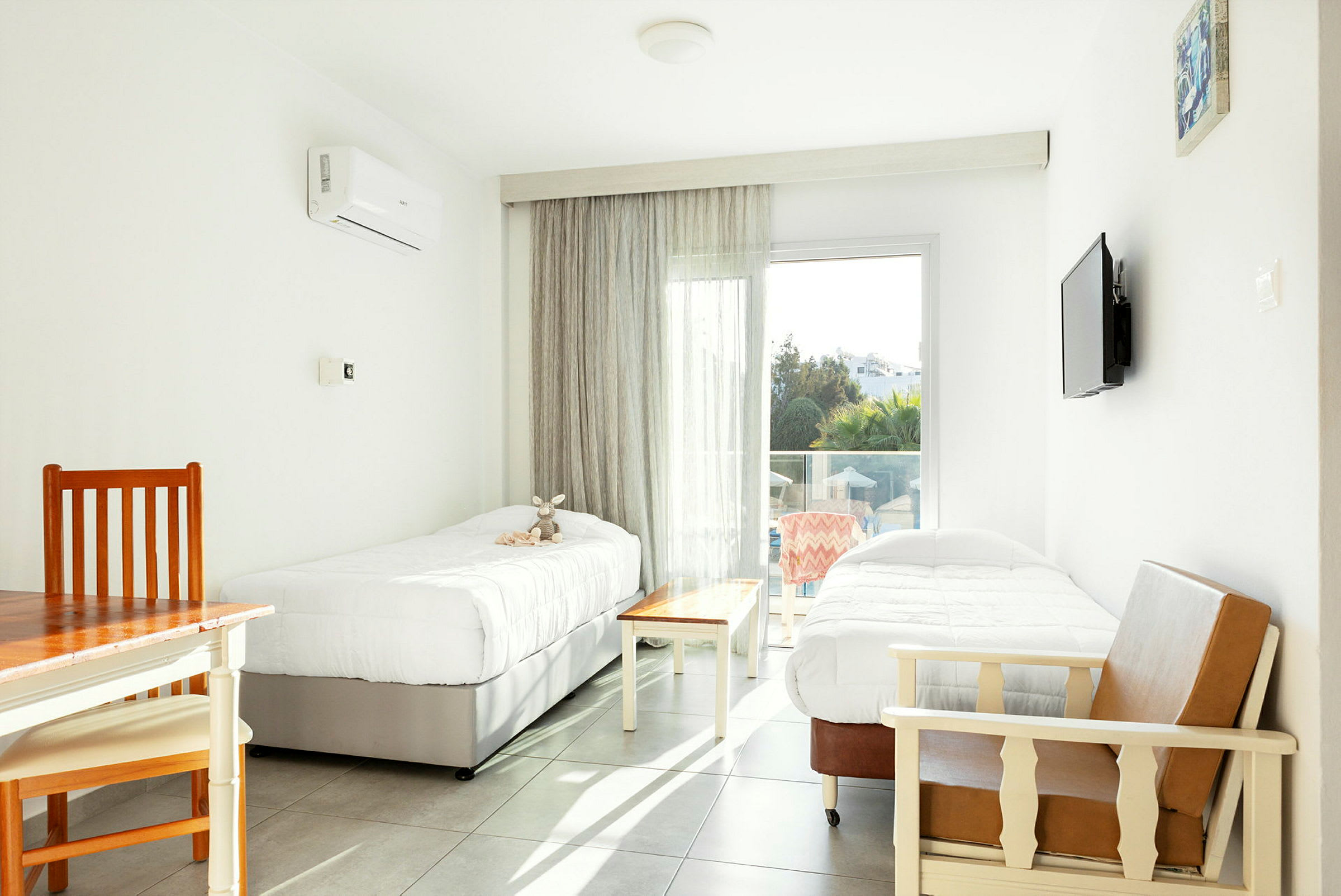 Ett ljust hotellrum med två enkelsängar, ett bord och stolar, samt en glasdörr som leder till en balkong. (AI-genererad text)