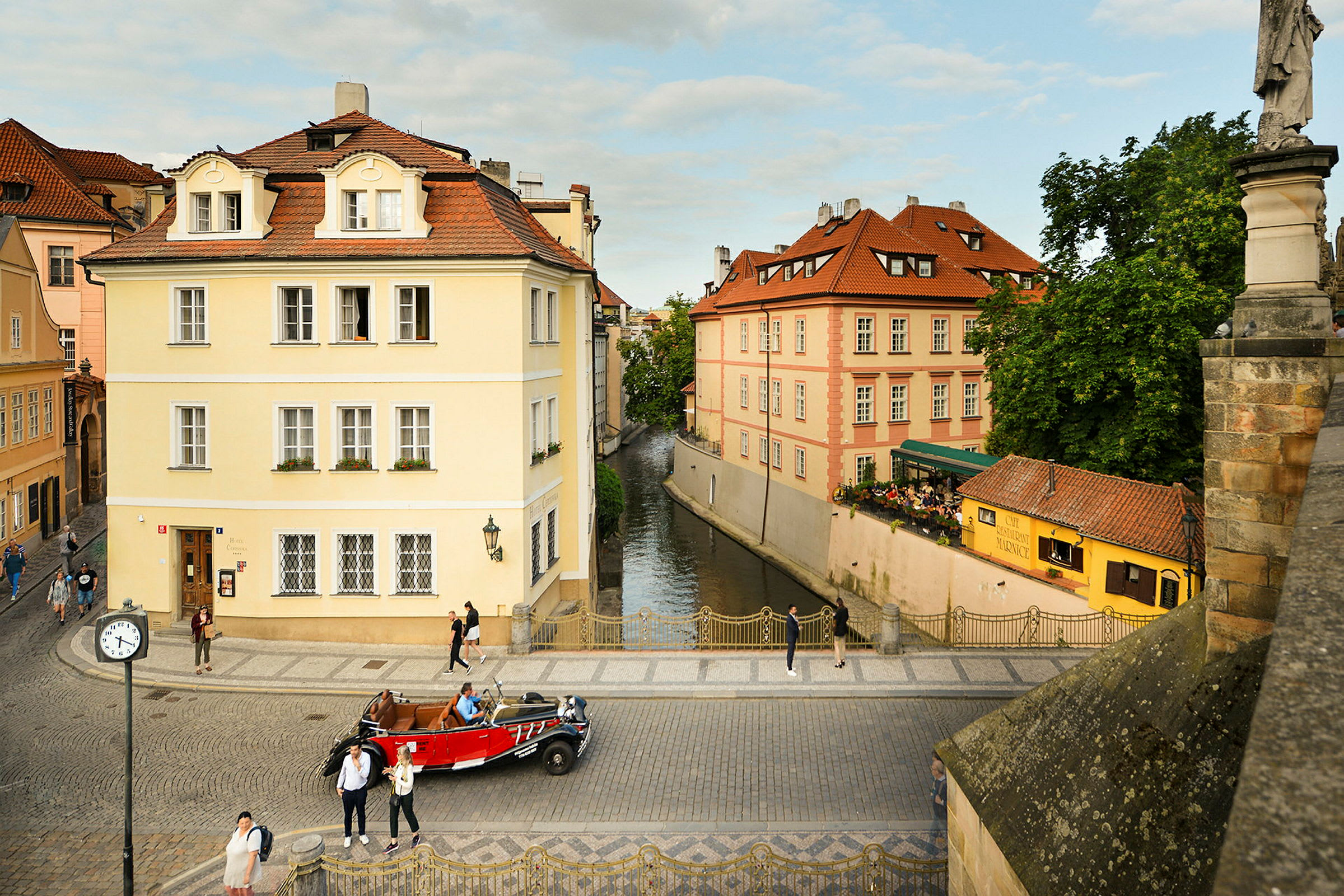 En gata med kullersten, flankerad av klassiska byggnader med färgglada fasader. En kanal syns mellan husen samt människor som går och en röd bil parkerad på gatan. (AI-genererad text)