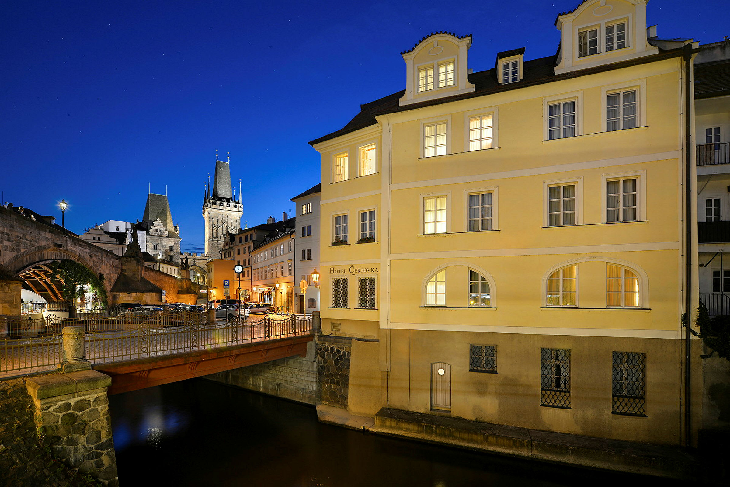 En liten bro över en kanal intill en gul byggnad under blå natthimmel. I bakgrunden syns en historisk stenbro och en kyrktornsliknande byggnad. (AI-genererad text)