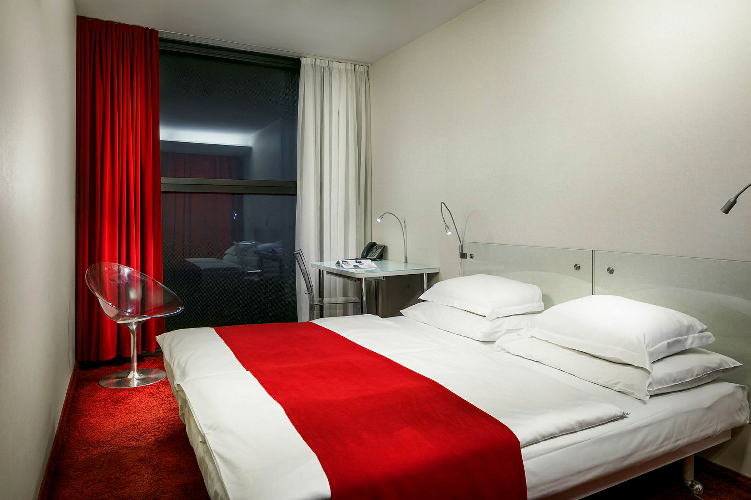 Hotellrum med röd matta, vit säng med rött överkast, glasstol och skrivbord. (AI-genererad text)