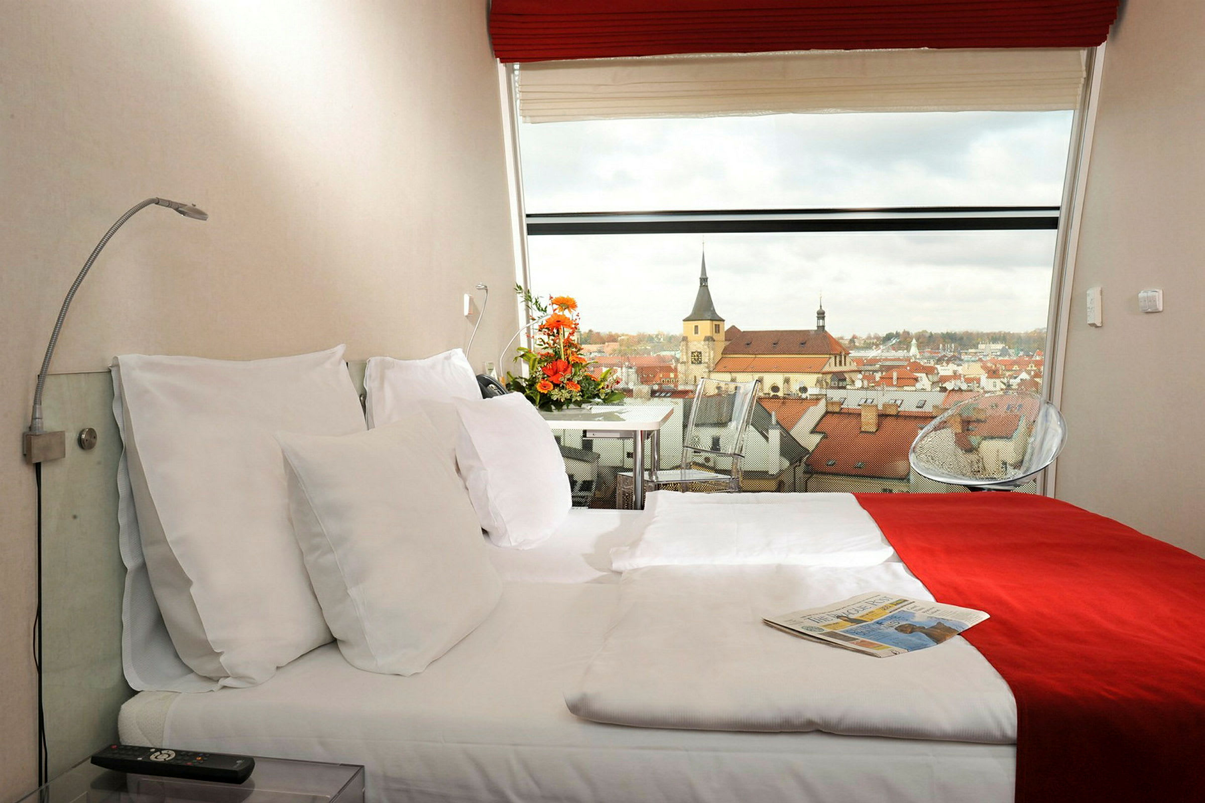 Ett hotellrum med en dubbelsäng, dekorerad med röda och vita sängkläder. En panoramautsikt över en stad med kyrkbyggnader syns genom fönstret. (AI-genererad text)
