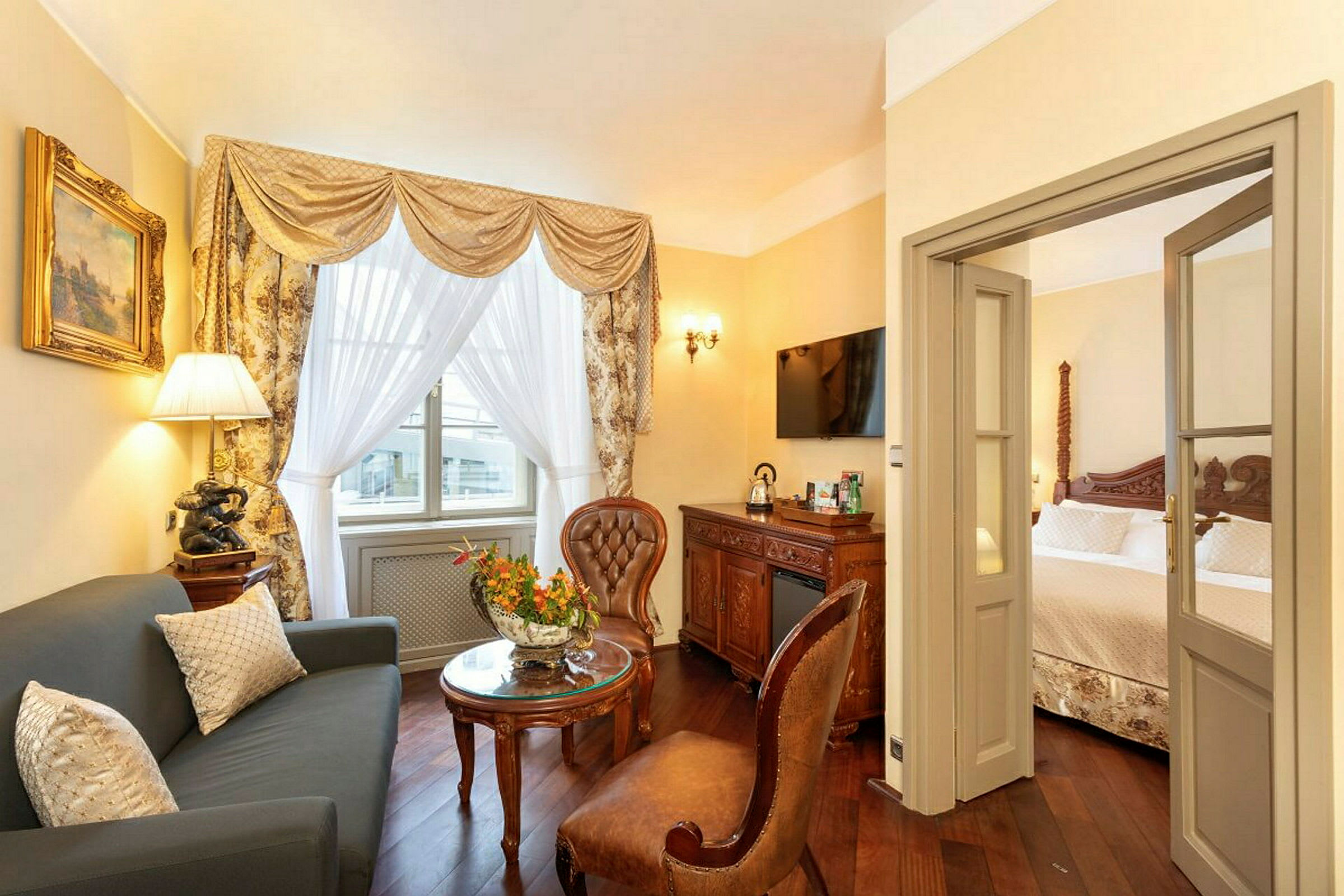 Ett elegant inrett hotellrum med ett vardagsrum med soffa och stolar samt ett sovrum i bakgrunden. Det finns en dekorativ gardin vid fönstret och en dörröppning mellan rummen. (AI-genererad text)