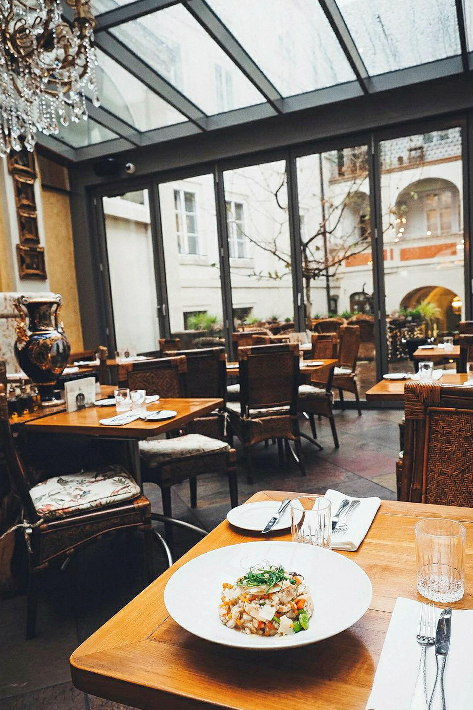 Ett restaurangbord är dukat med en tallrik mat och bestick, beläget i ett stilfullt inrett rum med stora glasfönster som ger utsikt till en innergård. (AI-genererad text)