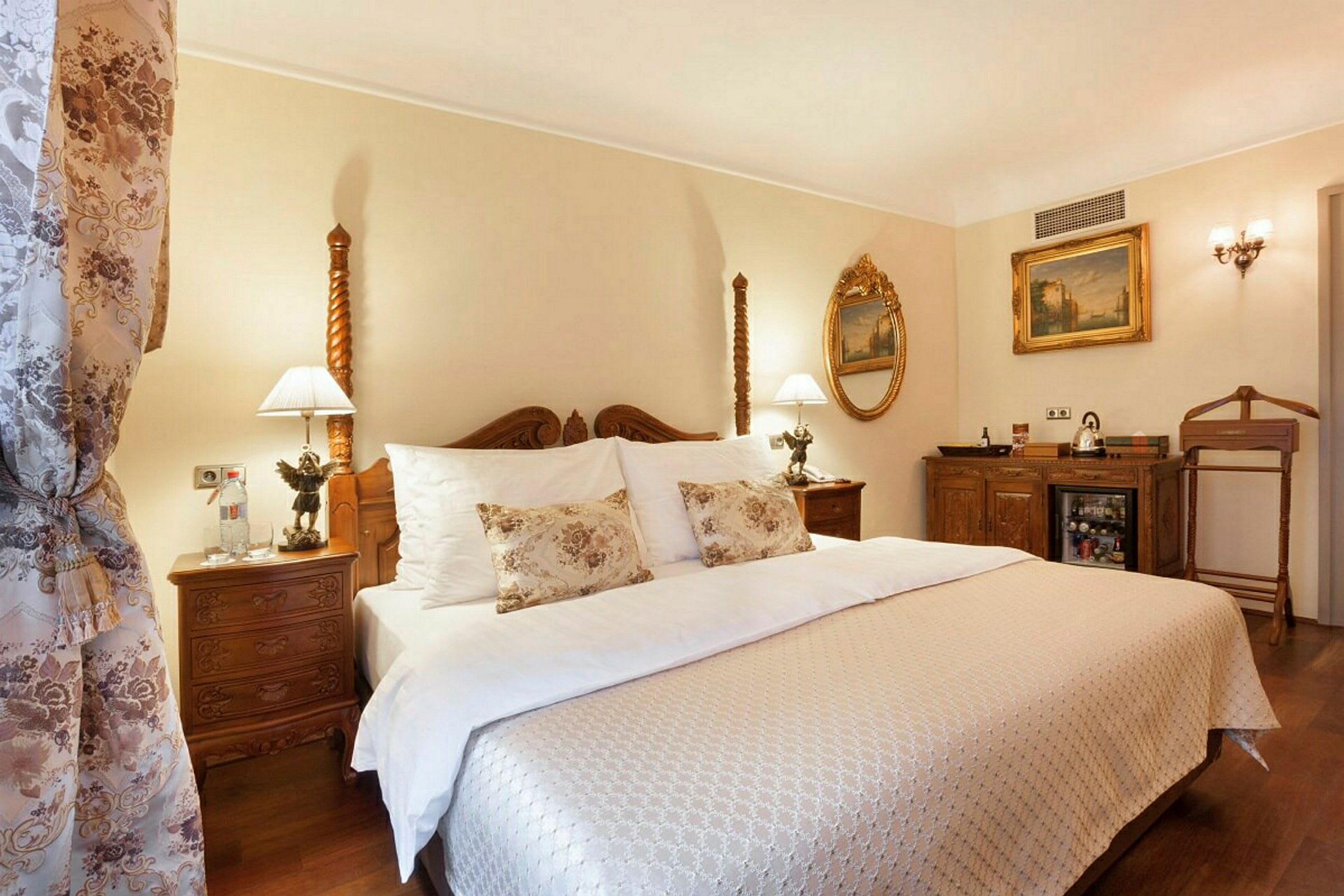 Ett elegant hotellrum med en dubbelsäng klädd i vita och mönstrade textilier, dekorationer i klassisk stil och lampor på sängborden. (AI-genererad text)