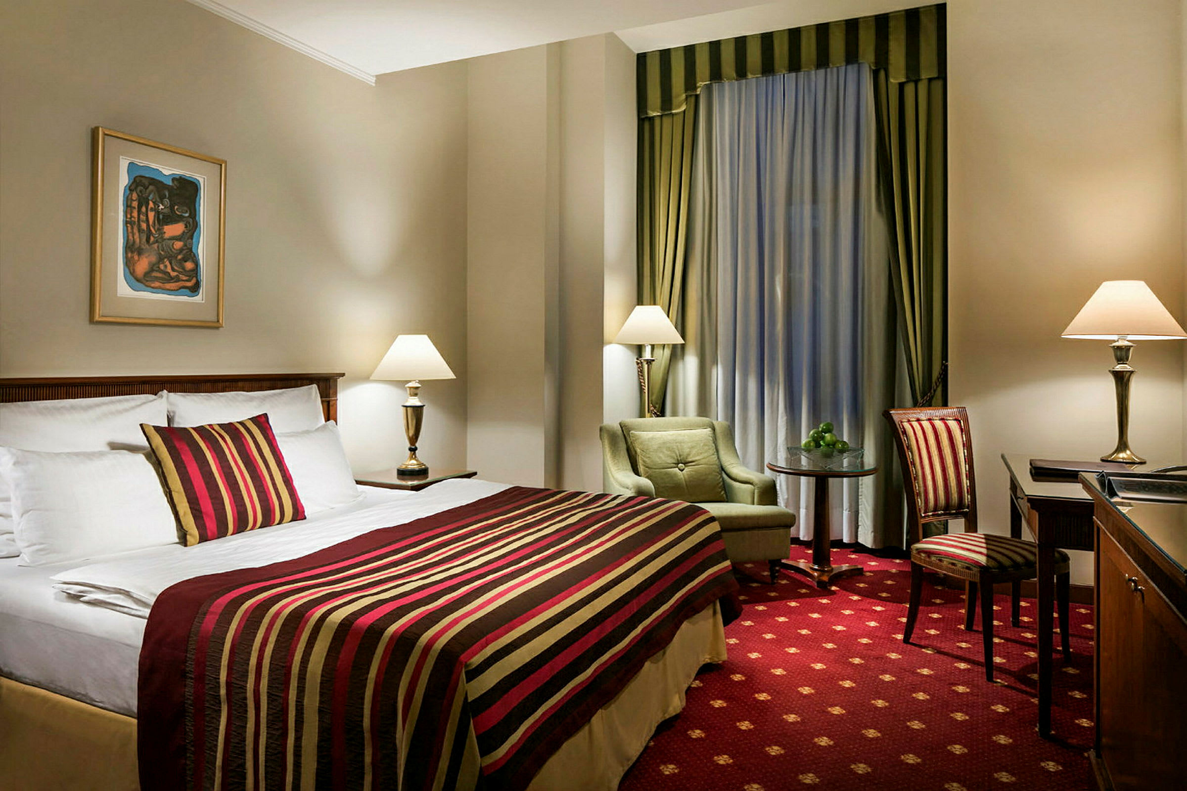 Ett hotellrum med en stor säng klädd i randigt överkast och prydnadskuddar. Det finns två bordslampor, en fåtölj med ett sidobord samt en stol vid ett skrivbord. Rummet är dekorerat med gardiner och en tavla på väggen. (AI-genererad text)