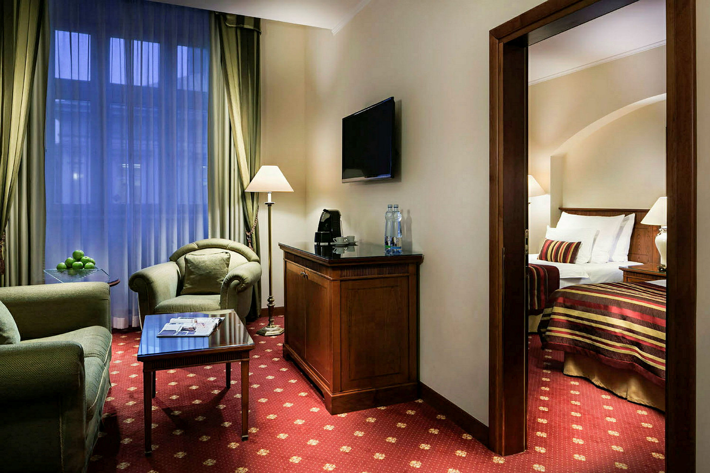Ett hotellrum med sittgrupp med soffa och fåtölj, ett soffbord med tidningar, och ett öppet sovrum med säng i bakgrunden. (AI-genererad text)