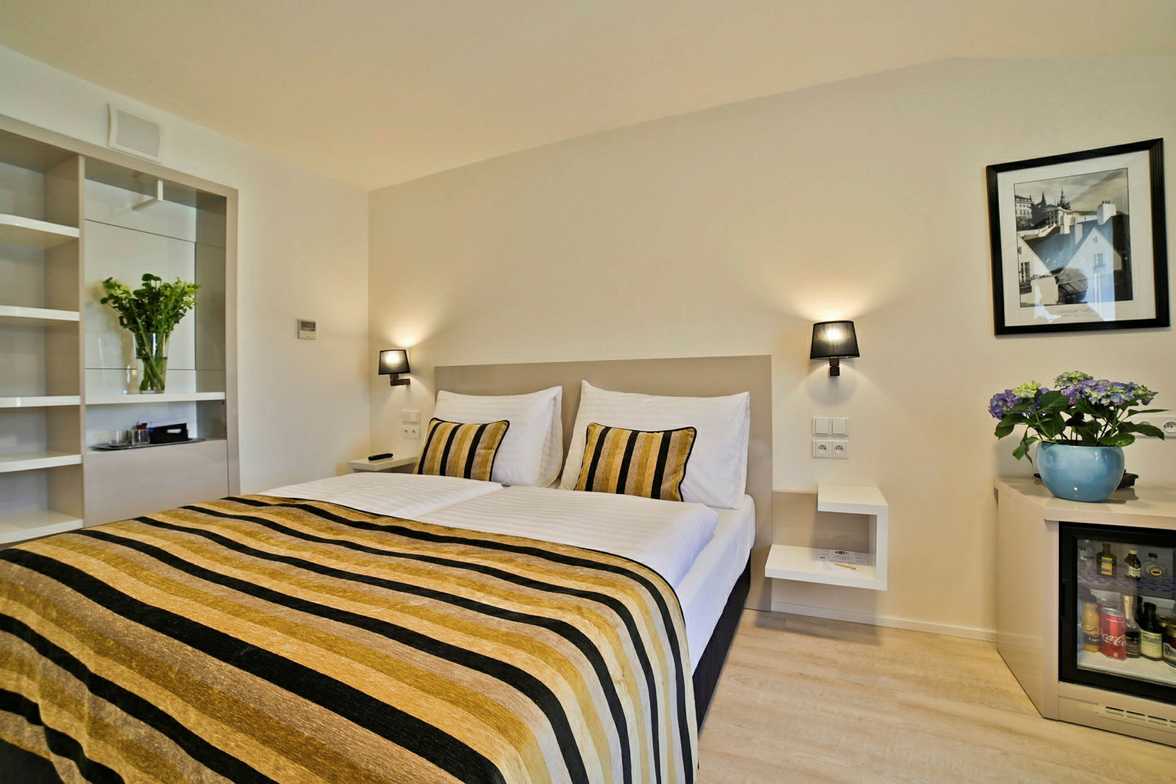 Hotellrum med dubbelsäng, randig sängöverkast i guld och svart, vita väggar, en växt och tavla på väggen samt ett litet kylskåp. (AI-genererad text)