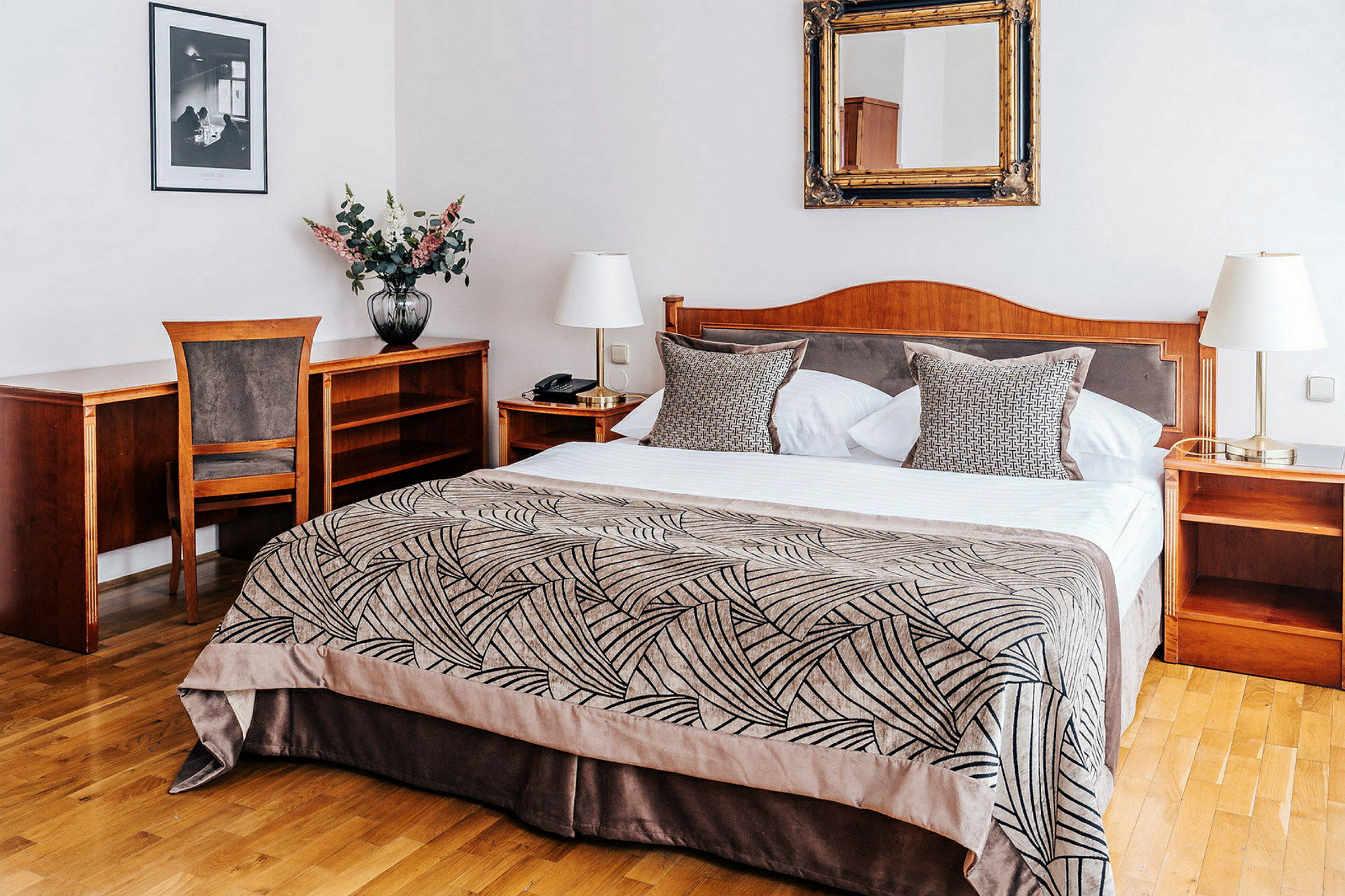 Ett hotellrum med en dubbelsäng, sängbord och skrivbord av trä. På sängen ligger ett mönstrat överkast och kuddar. (AI-genererad text)